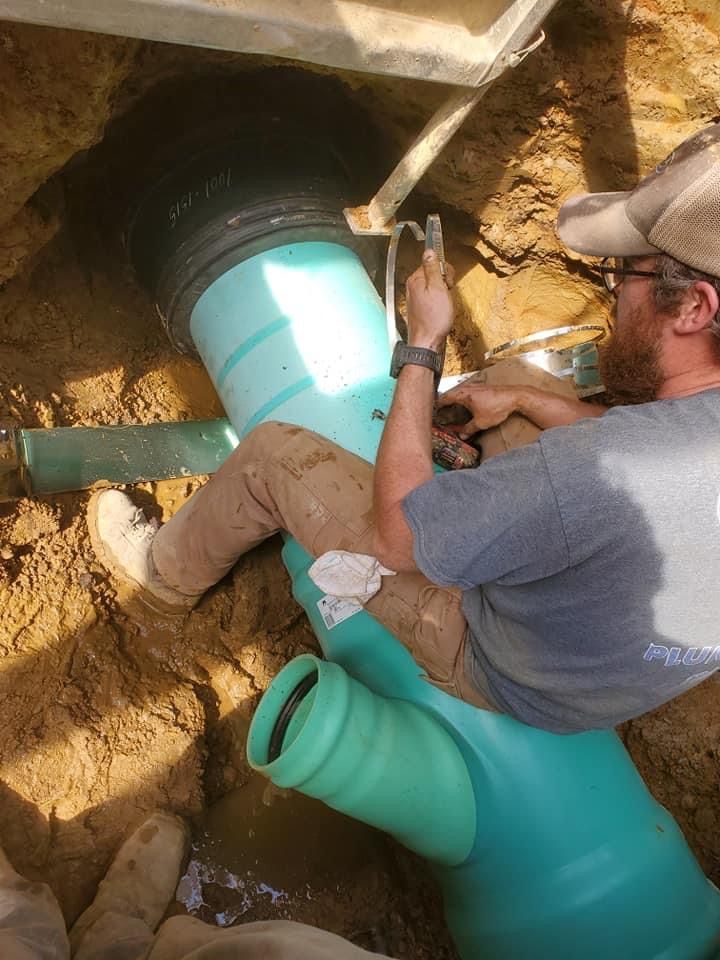 Man in work clothes connecting green pipes in a dirt trench, using a tool.