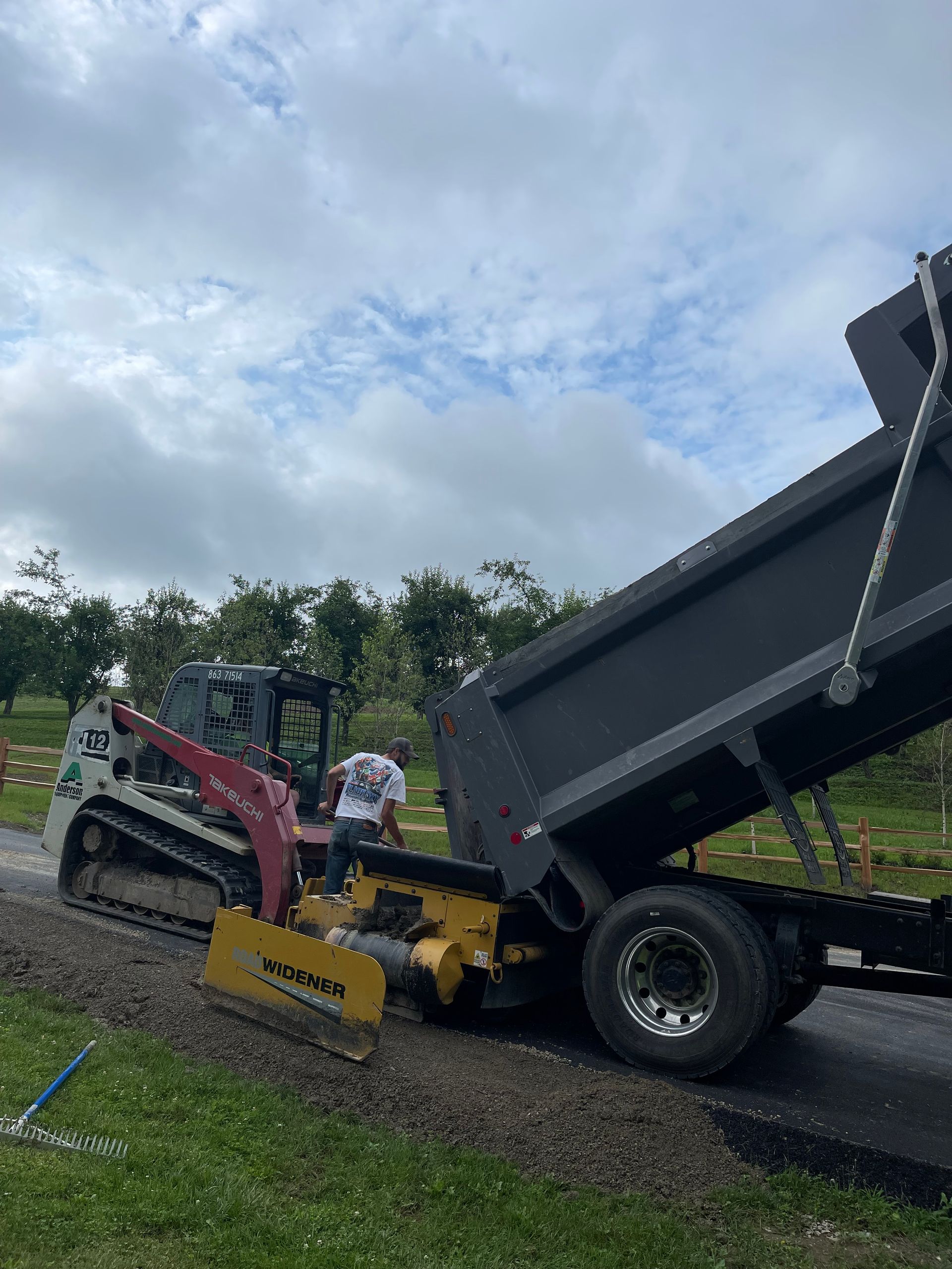 A dump truck unloading gravel next to a paving machine on a construction site.