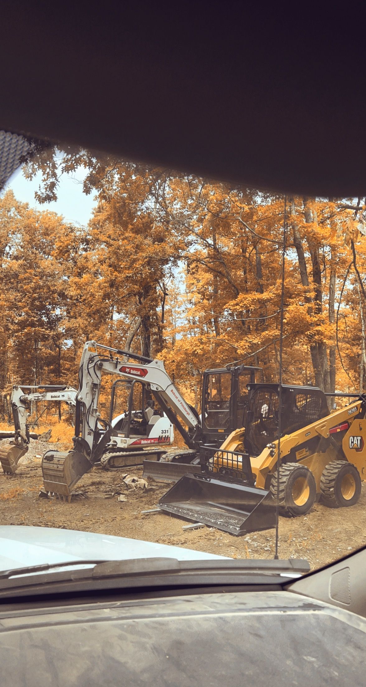 Yellow excavator and skid steer working in a wooded area with trees displaying orange leaves.