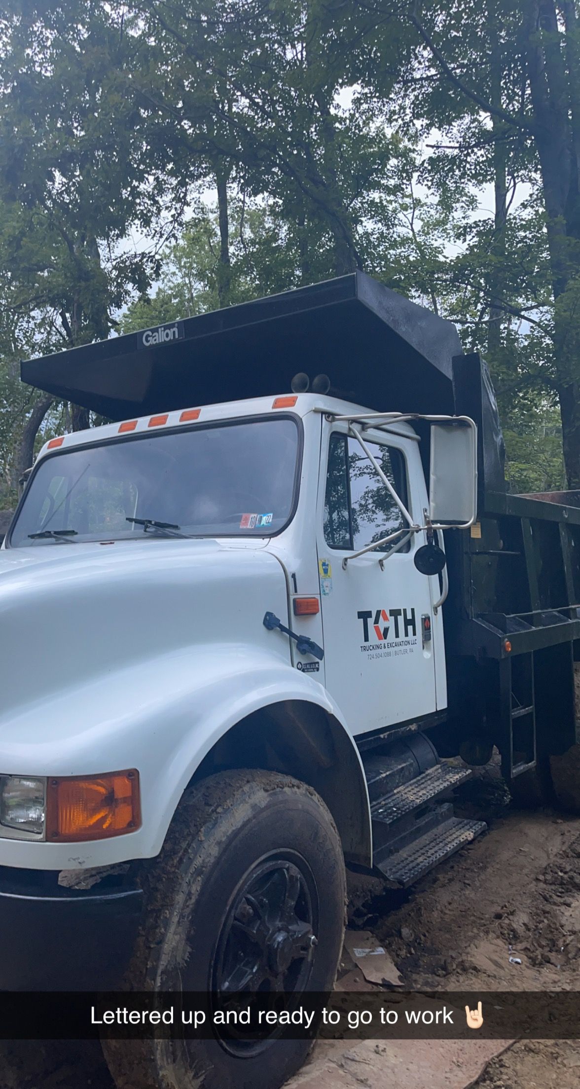 White dump truck parked on a dirt road, trees in the background, labeled 