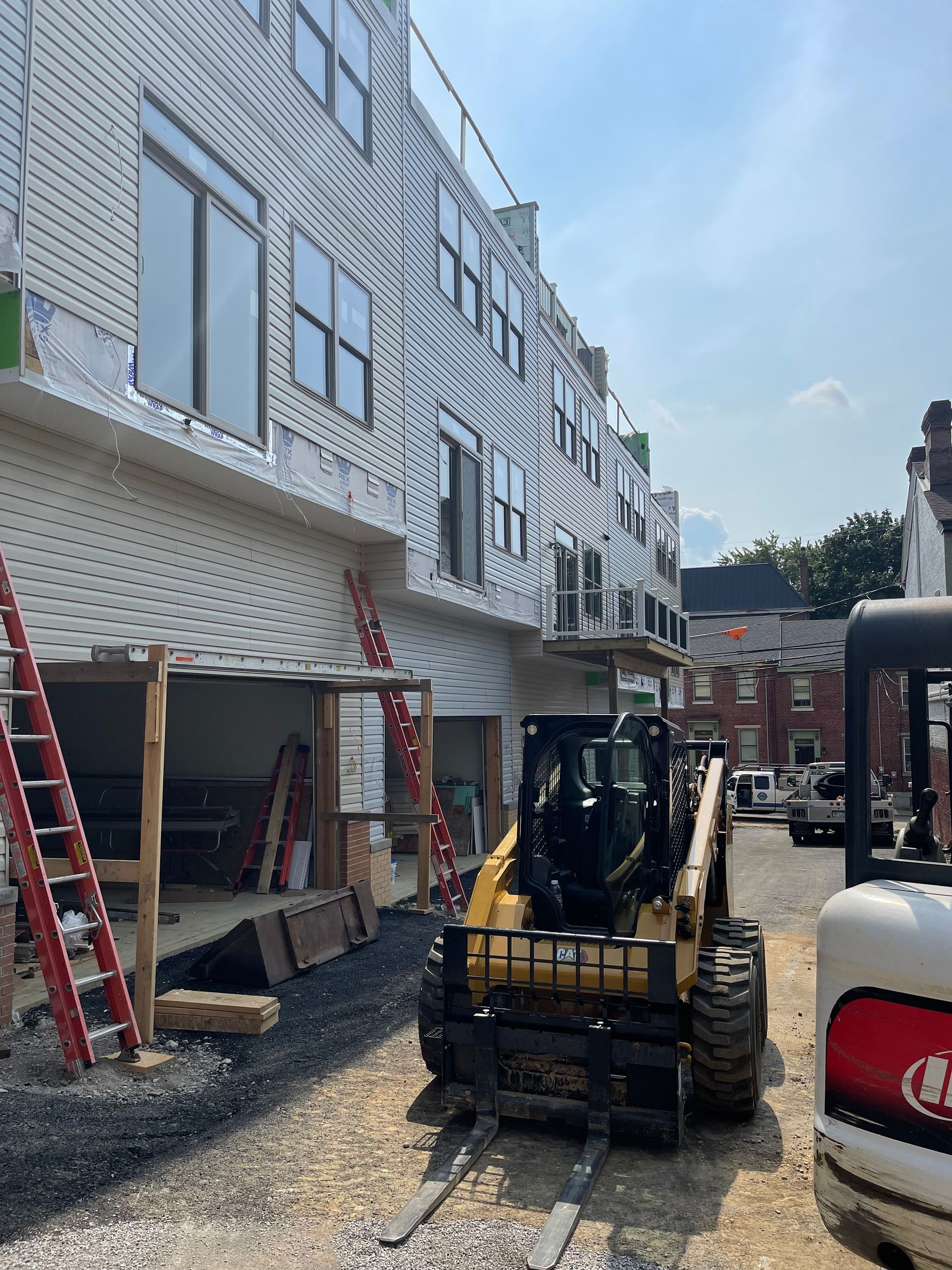 Construction site: Yellow skid steer, building with white siding, ladders, excavation, and a sunny sky.