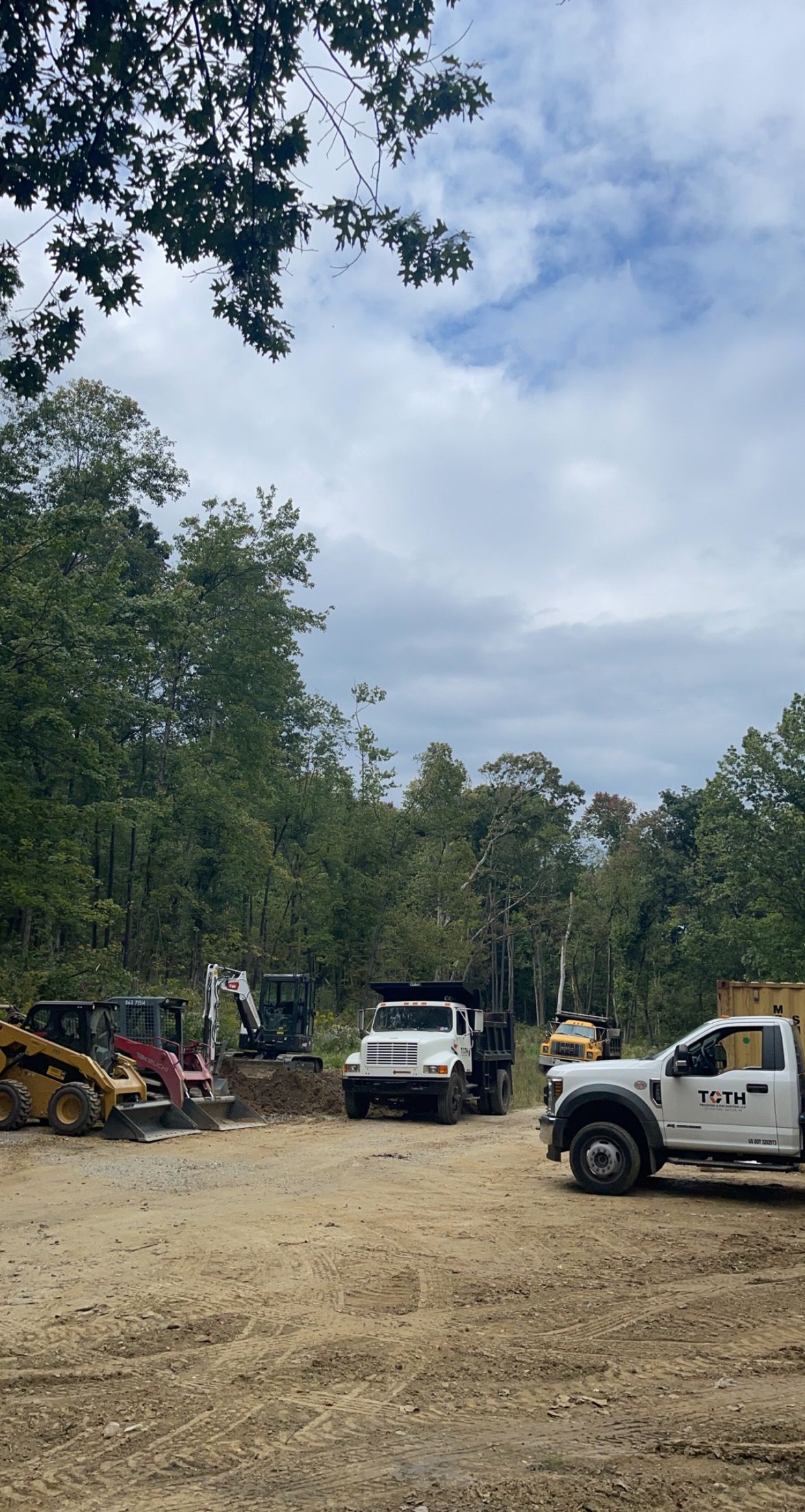 Construction site with dump trucks and heavy machinery in a wooded area under a cloudy sky.