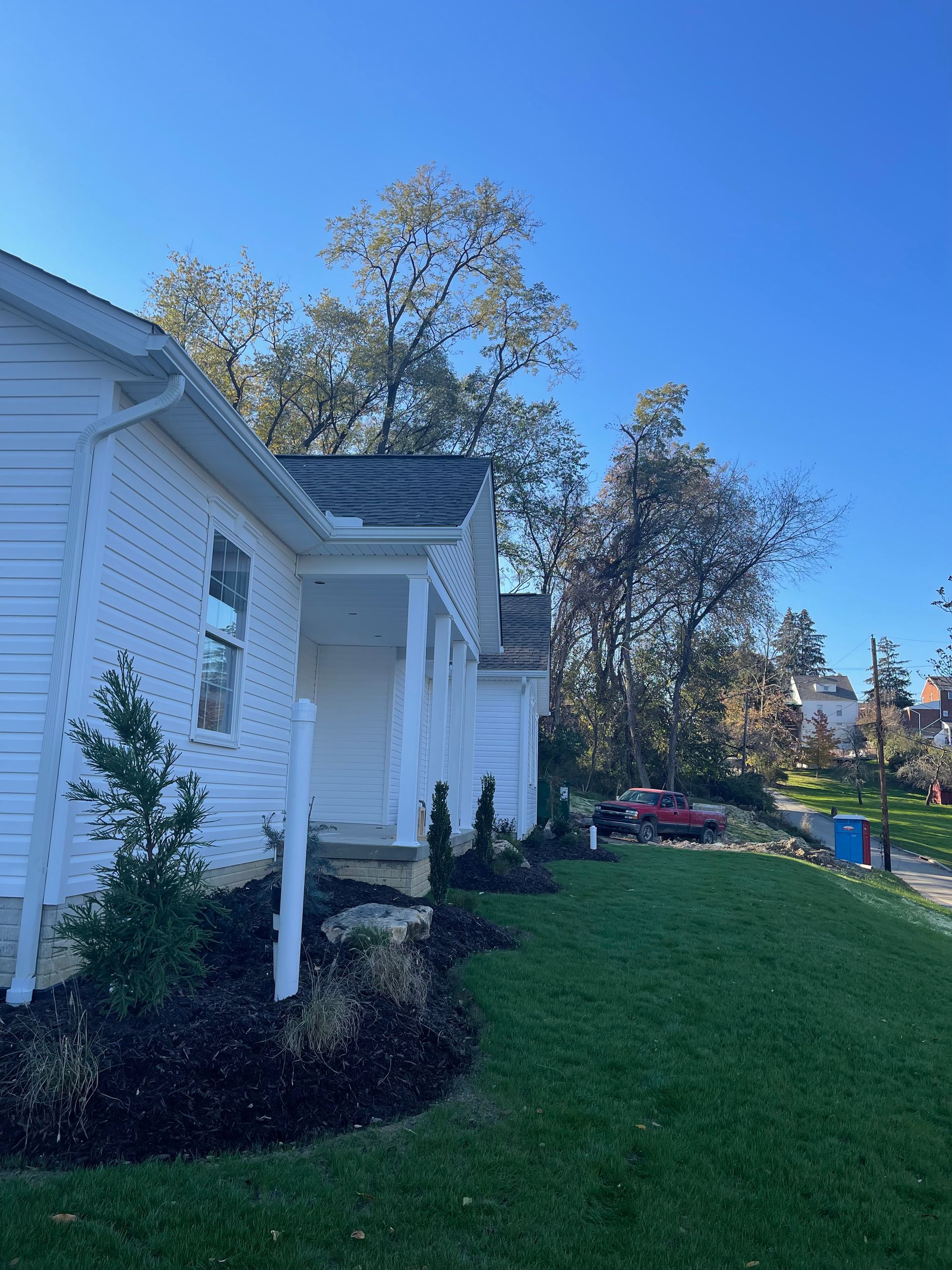 White house with a small porch, green lawn, trees, and a blue sky on a sunny day.