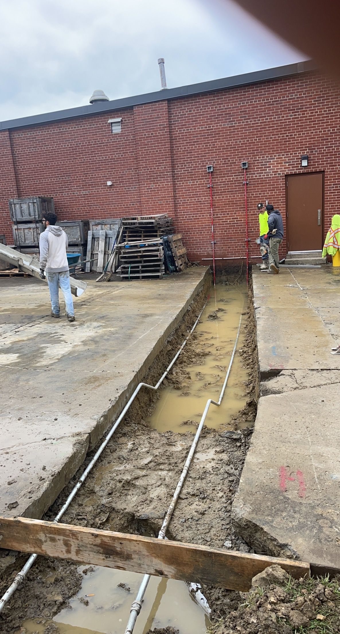 Workers excavate a trench along a corrugated metal building, containing pipes and standing water.