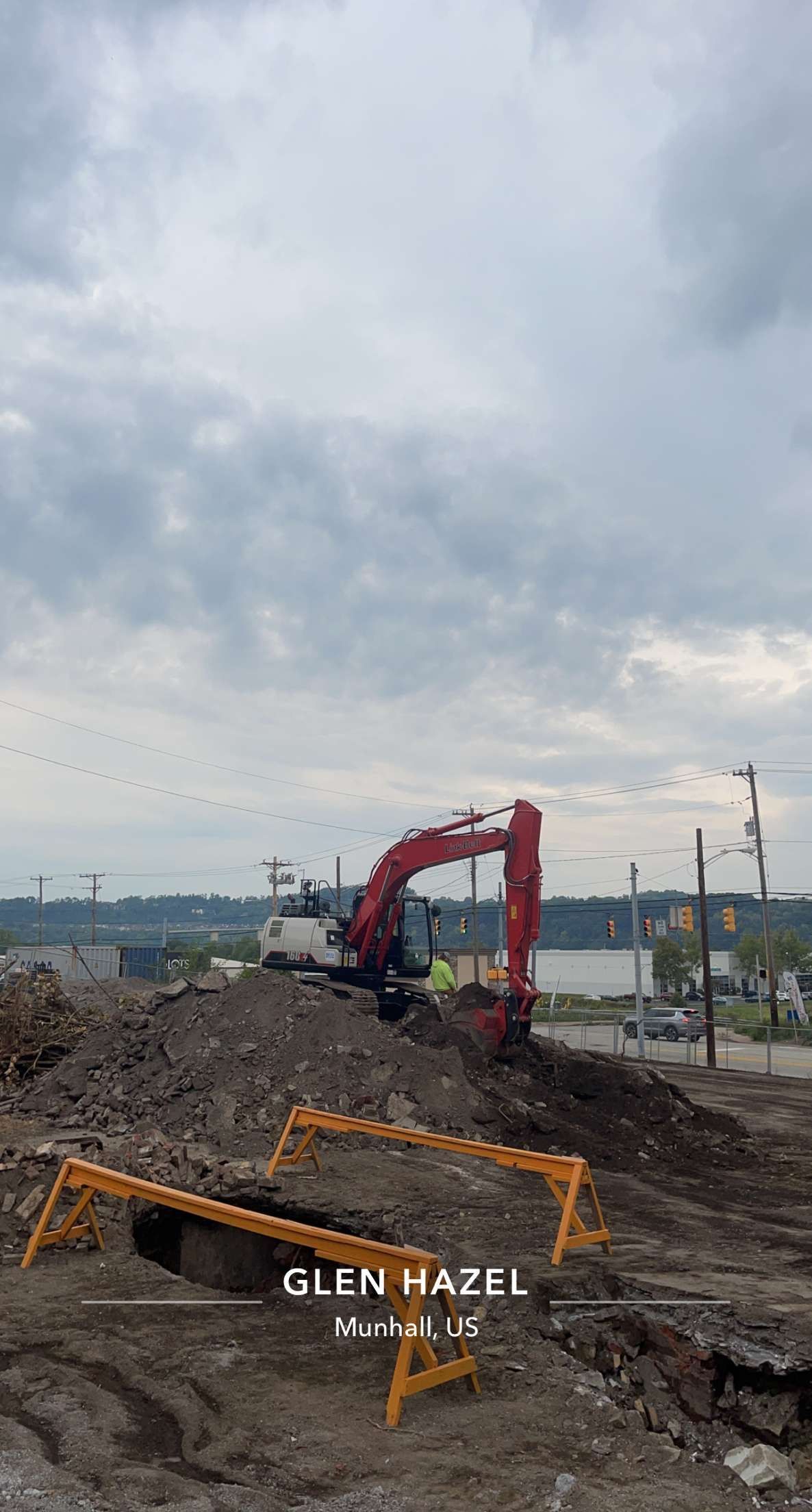 Red excavator on a dirt pile under a cloudy sky, with yellow safety barricades in the foreground.
