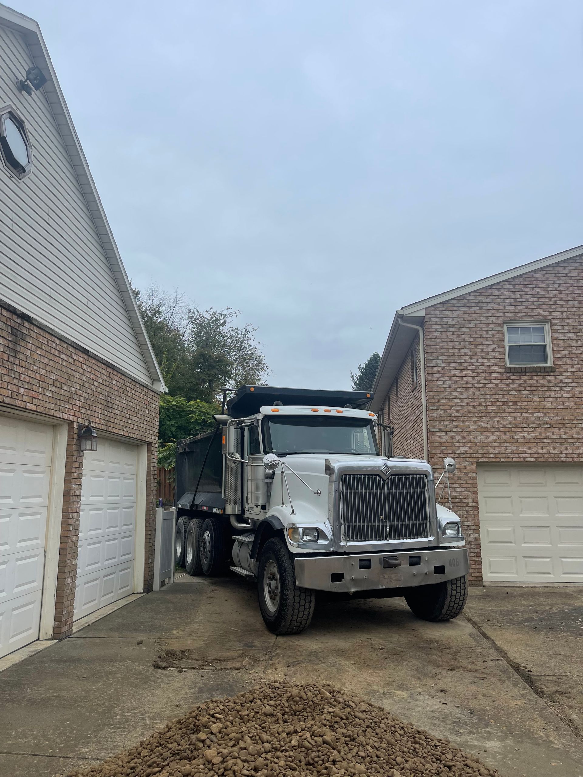 White dump truck parked between two brick buildings. Pile of rocks in front. Overcast sky.