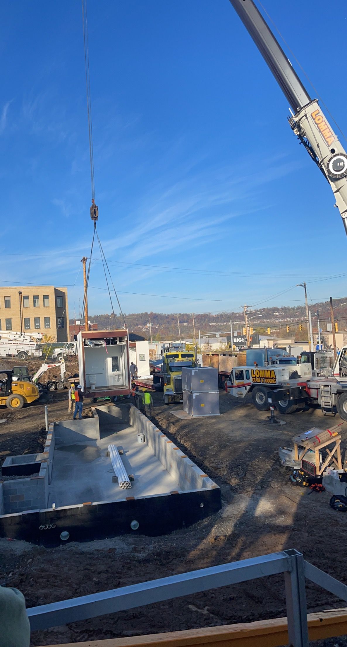 Crane lifting building components at a construction site. Gray and white structures, blue sky, heavy machinery.