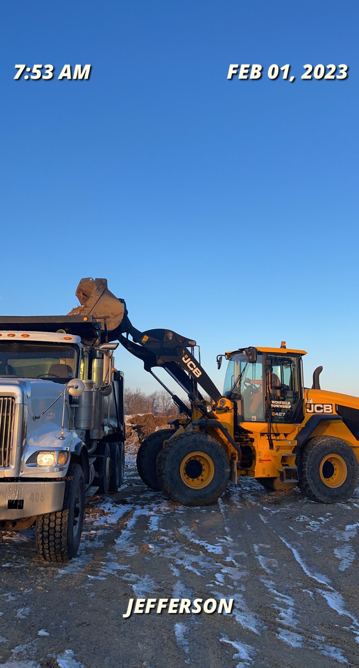 A yellow loader fills a dump truck with dirt on a cold, sunny day.