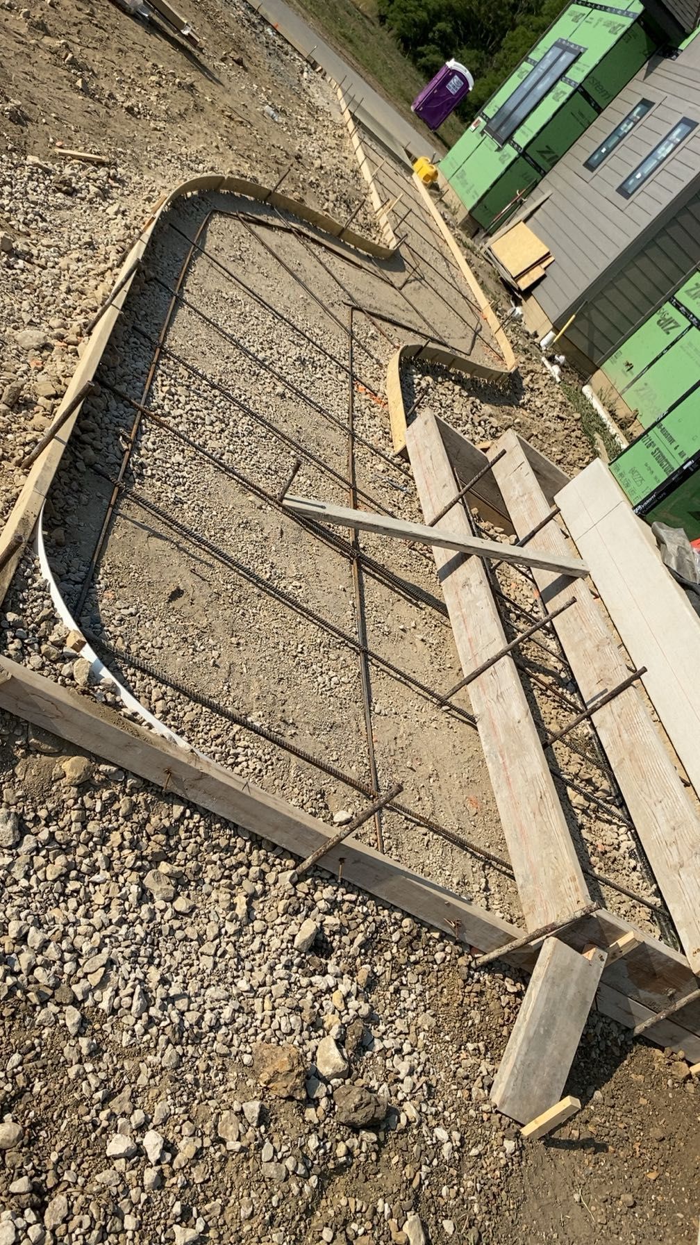 Construction site: concrete forms with rebar for a sidewalk. Wooden boards, gravel, and supplies nearby.