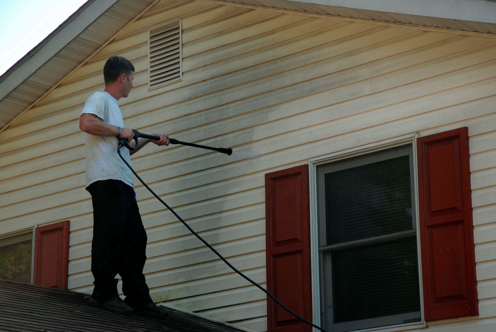Man pressure-washing a house roof and siding with a long sprayer wand