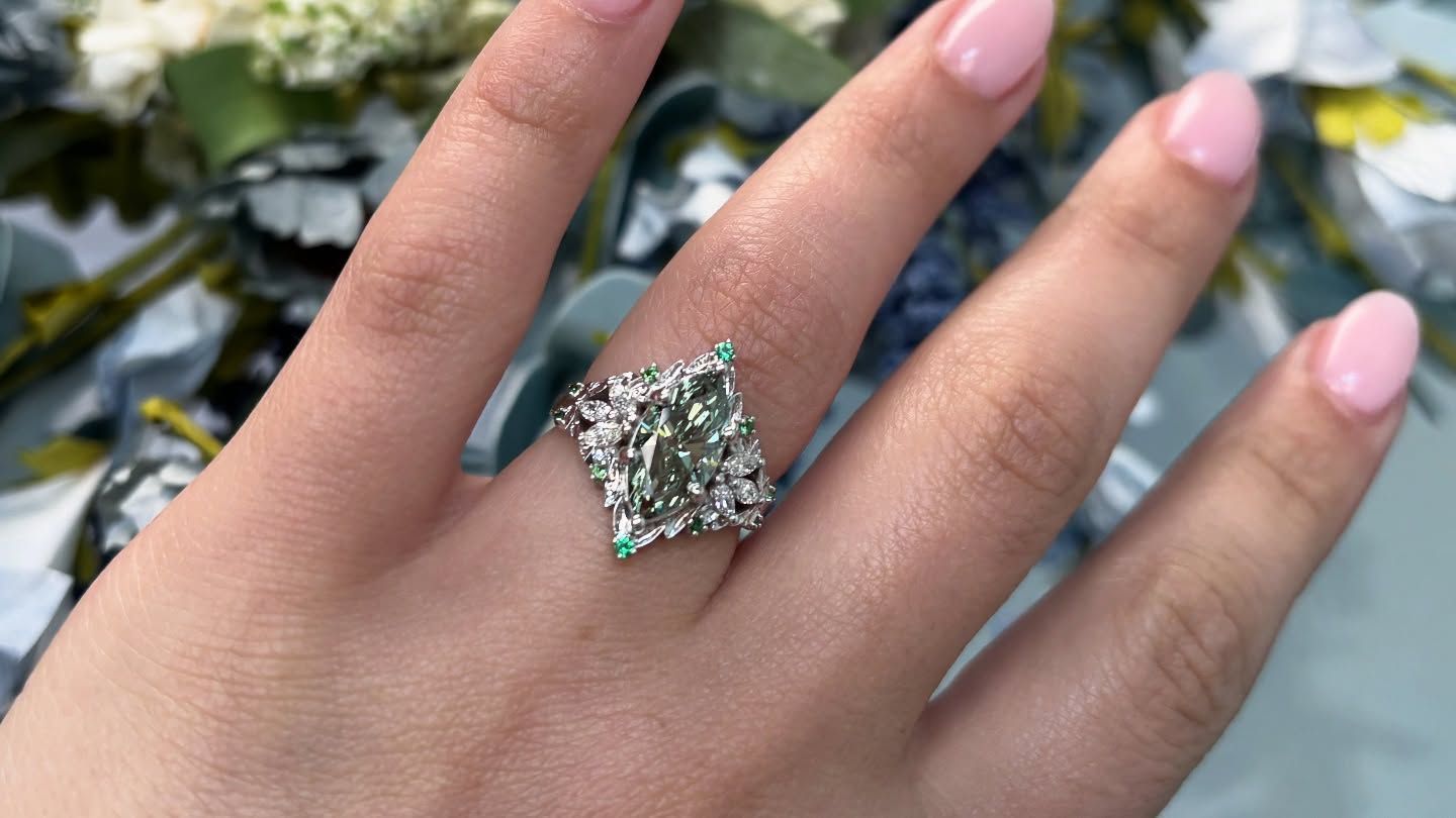 Hand wearing a large, sparkling gemstone ring with a pale pink manicure against a blurred garden background