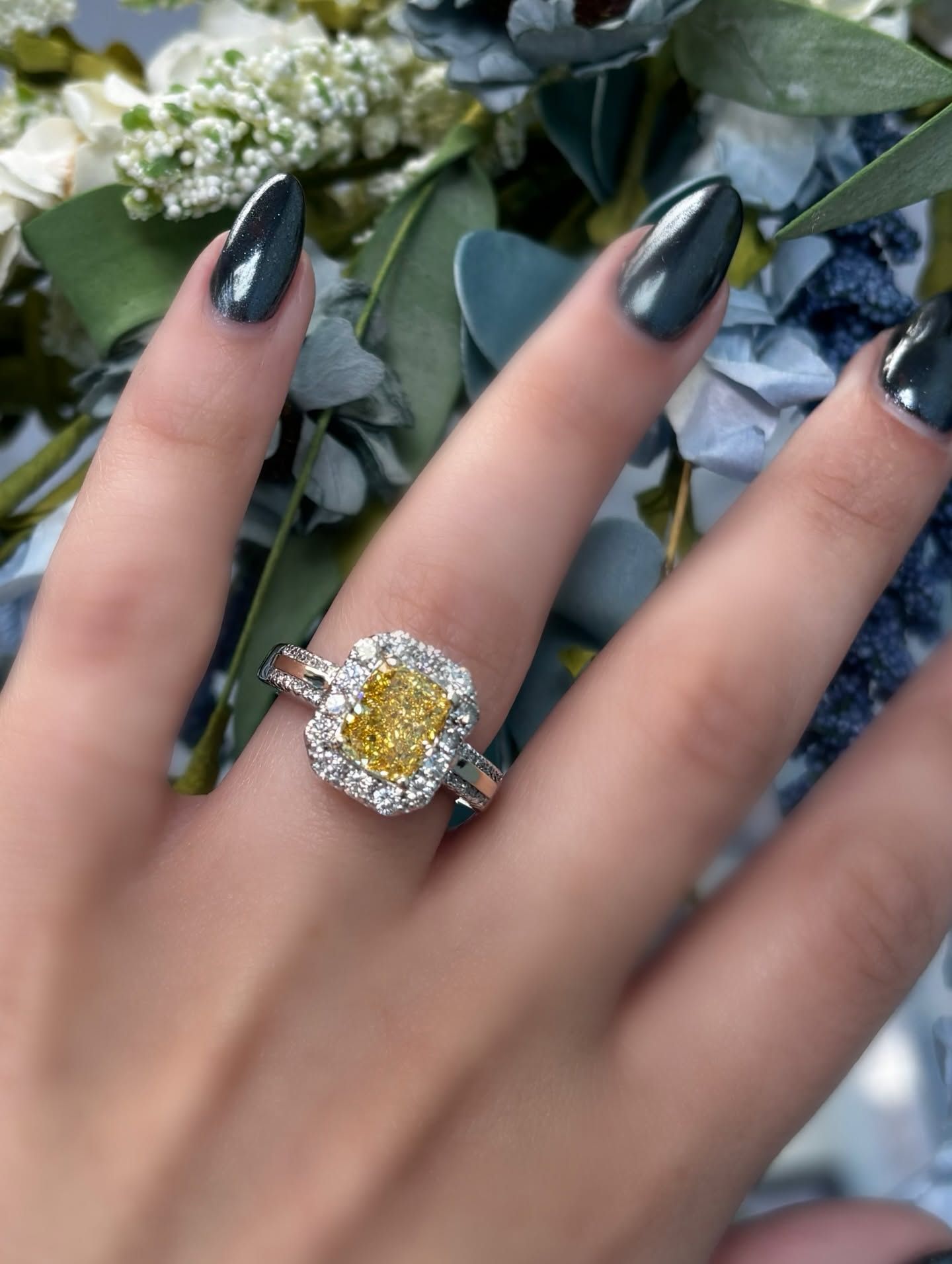 Hand with dark manicure wearing a yellow gemstone ring against blurred greenery and flowers