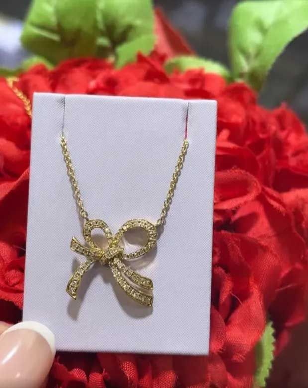 Gold bow-shaped necklace pendant on a white card, with red flowers in the background