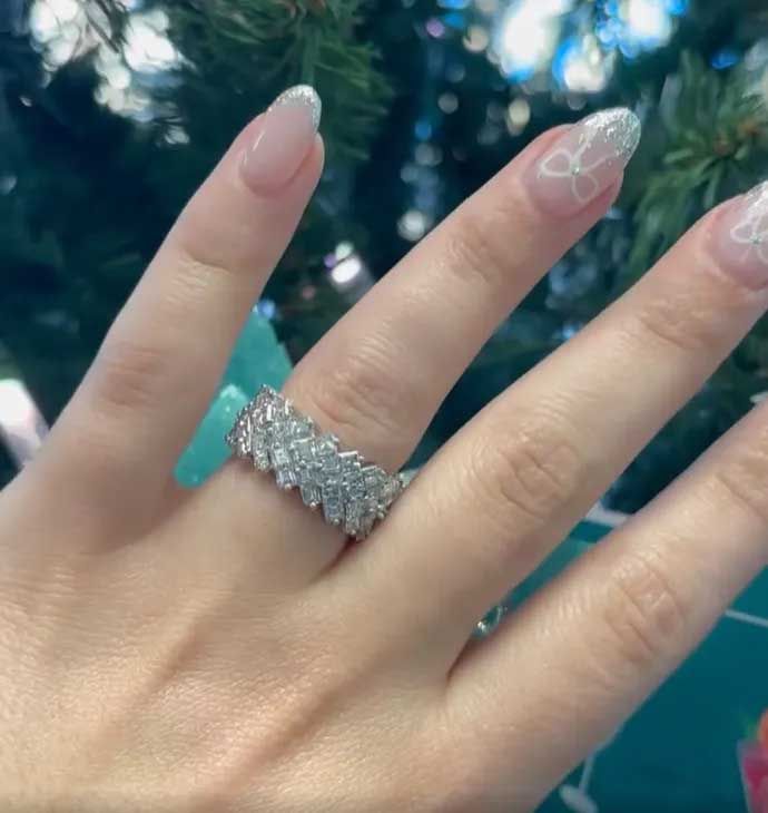 Hand with long nude nails wearing a sparkly silver ring against a blurred blue-green background