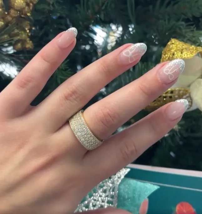 Hand with long nude-and-white lace nails wearing a sparkling gold ring near a decorated Christmas tree.