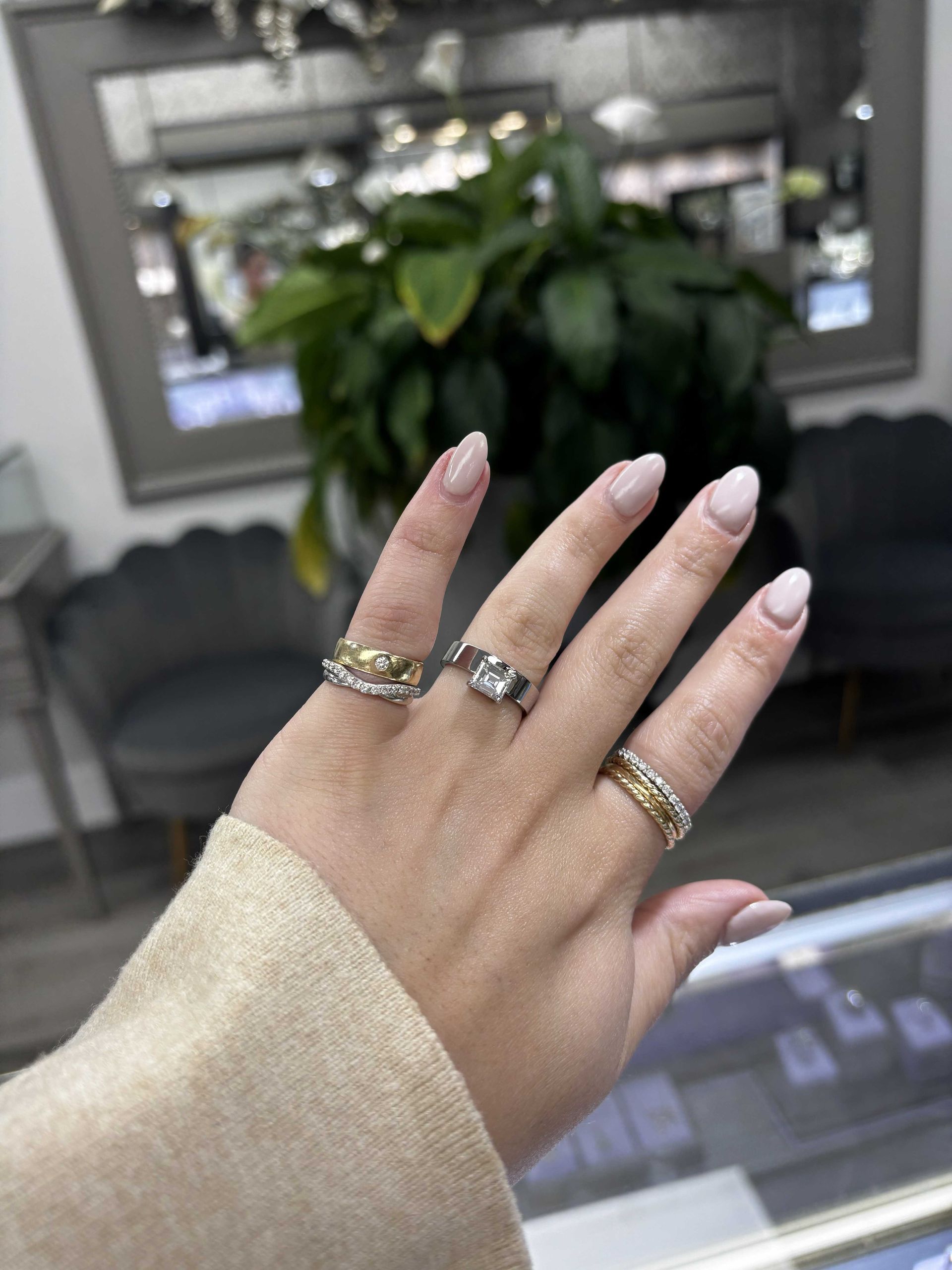Hand with pink nails and gold rings held up in front of a blurred plant and living room background