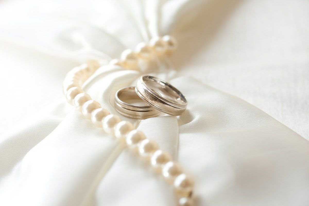 Two gold wedding rings resting on a white pearl necklace on white fabric