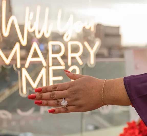 Hand with red nails and engagement ring reaching toward glowing “Will you MARRY ME” sign