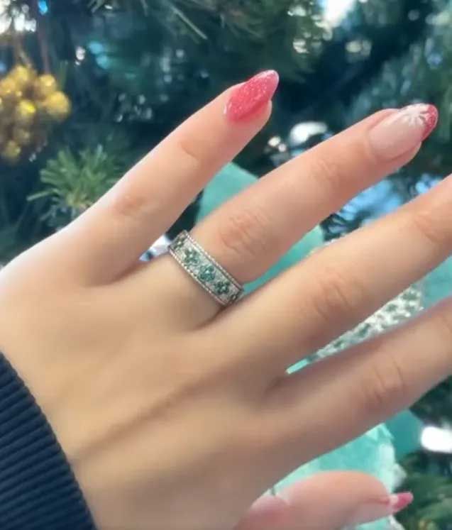 Hand with pink nails wearing a silver ring, held in front of a blurred Christmas tree.