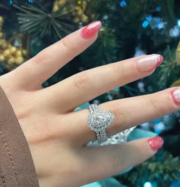 Hand with pink nails wearing a sparkling silver ring against a blurred evergreen background
