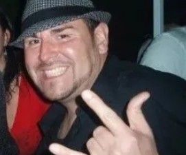 Smiling man in a black fedora flashes a hand gesture at a party
