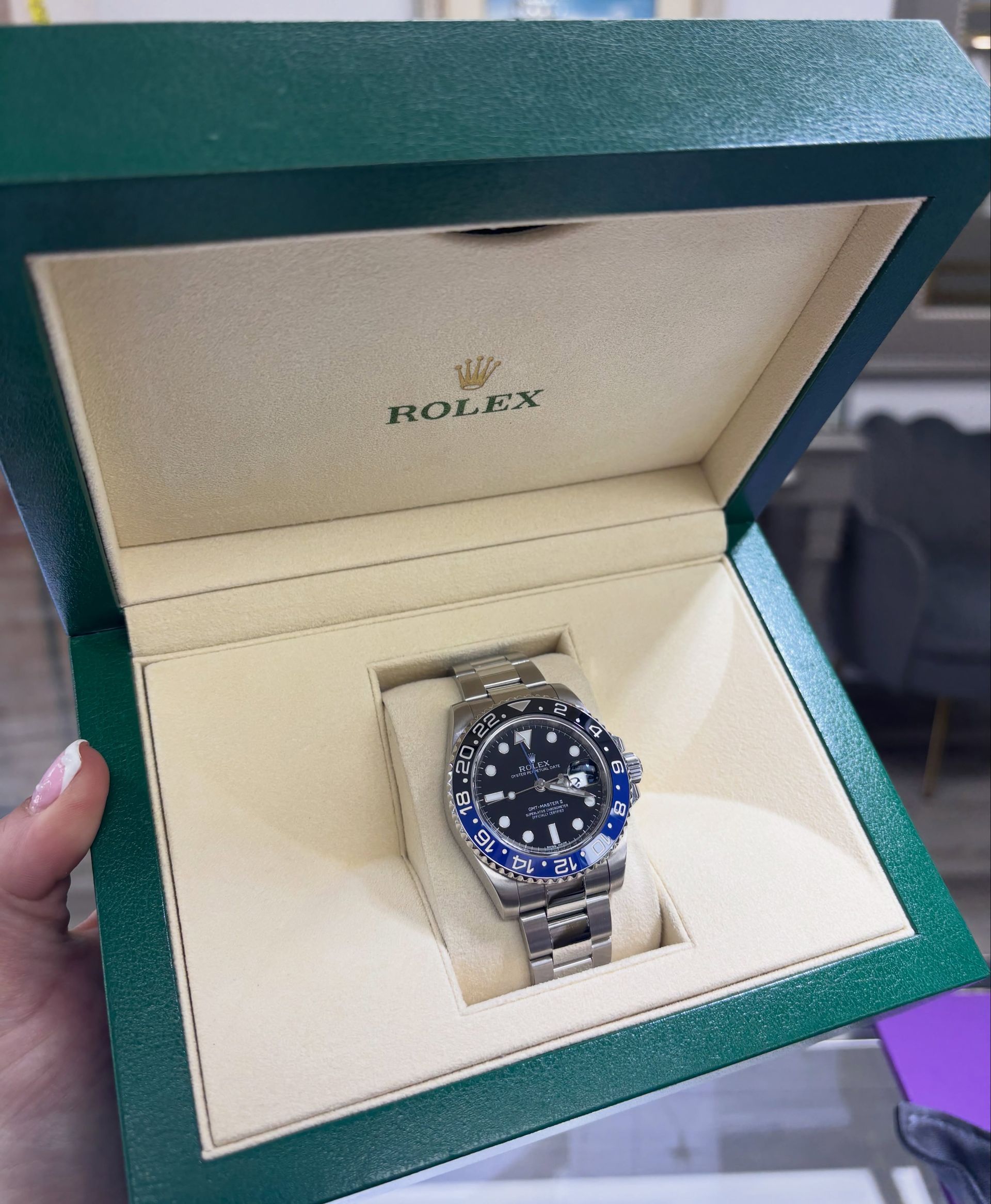 Rolex watch in a green presentation box, silver band and blue bezel, held open by a hand.