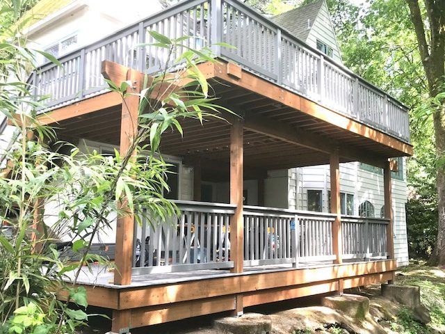 A large wooden deck with a gray railing is in front of a white house surrounded by trees.