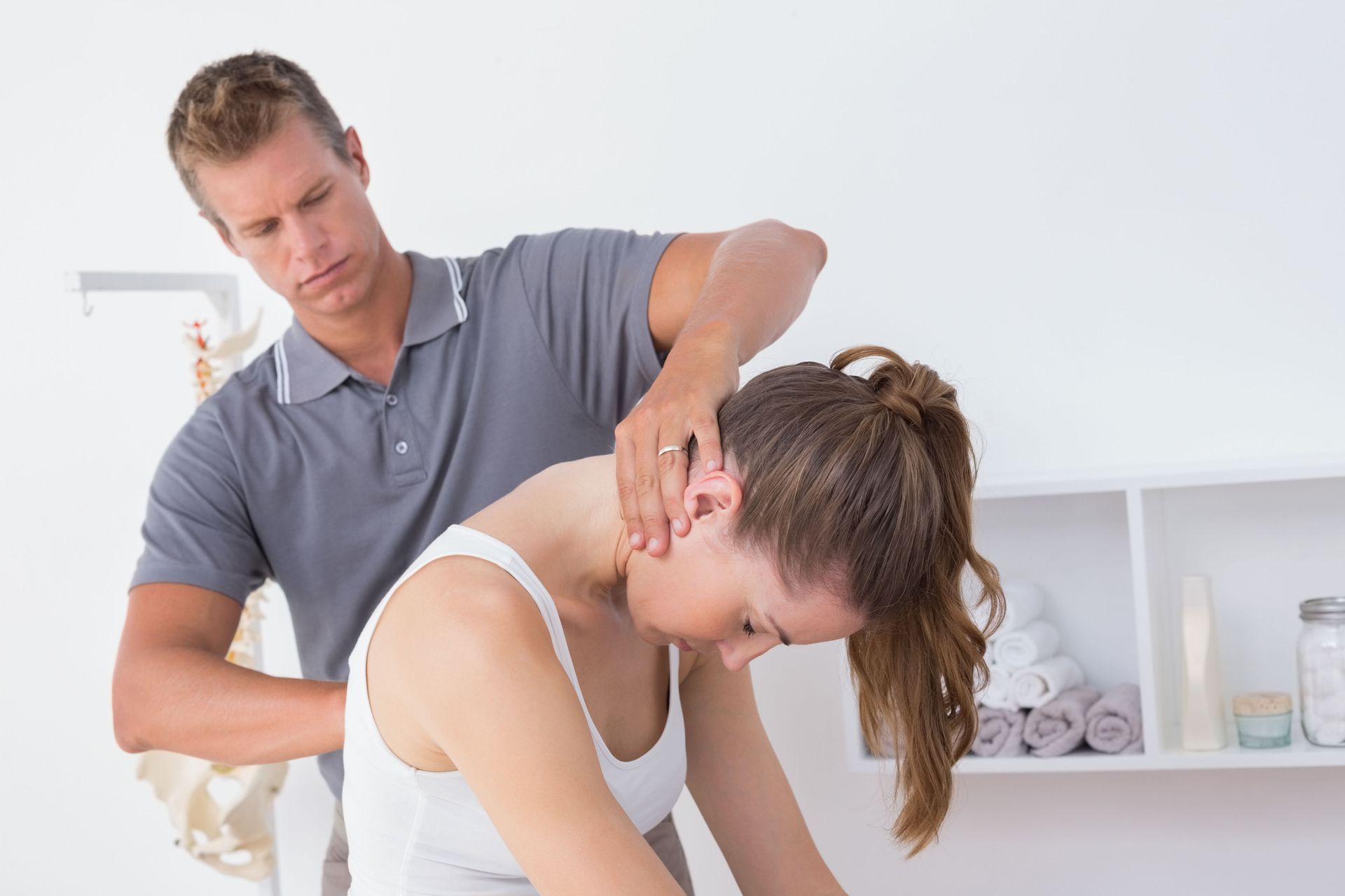 Chiropractor adjusting a woman's neck.  Both are in a light-filled room; the woman is bending forward.