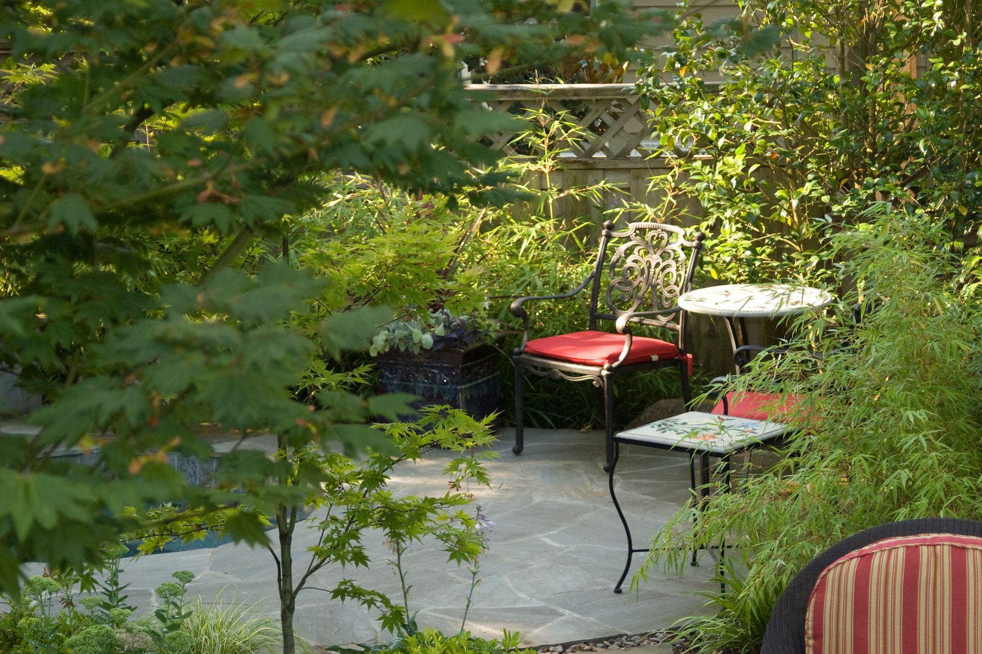 Garden patio with black chairs, a red-cushioned seat, and a small round table amid lush greenery