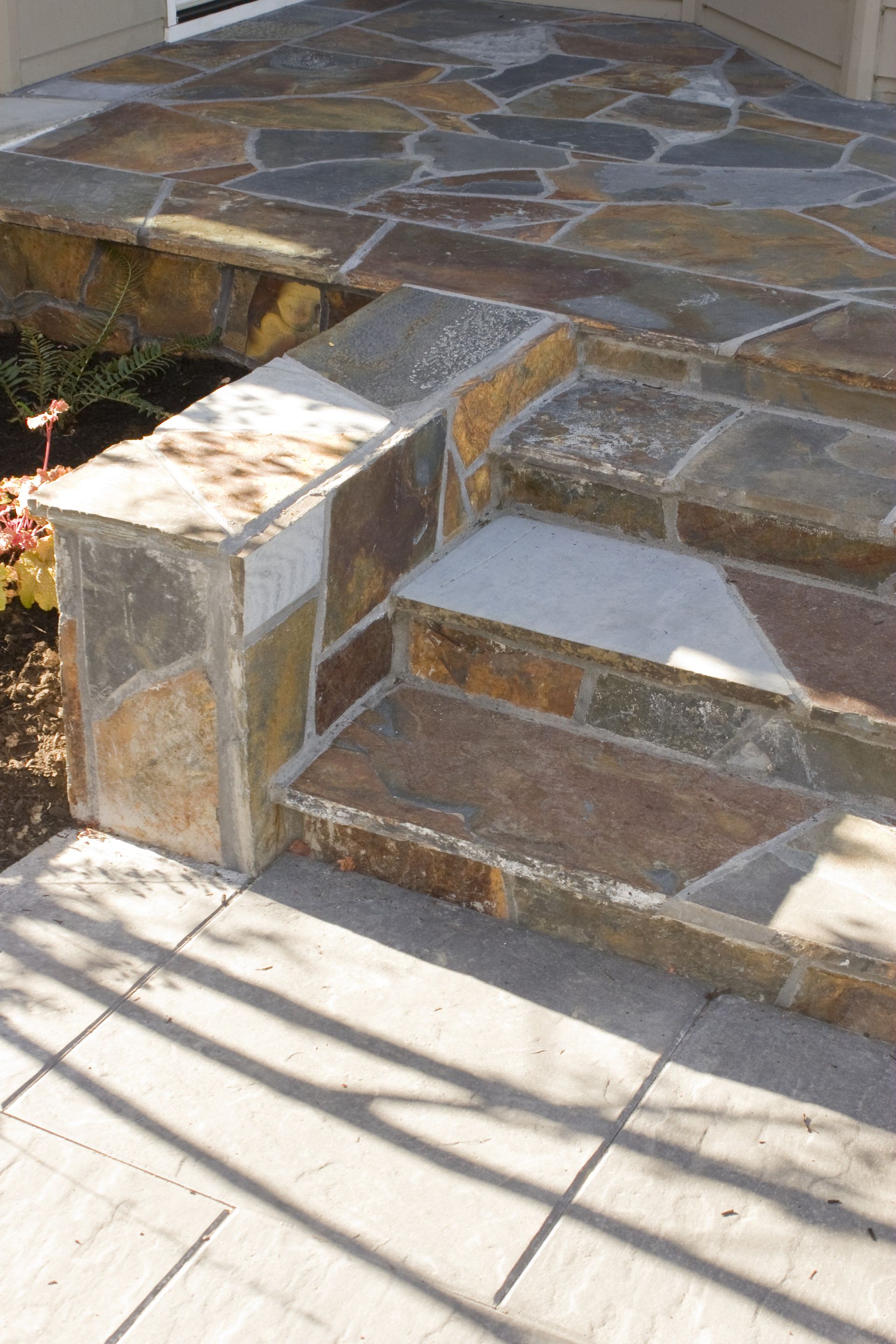 Stone steps and a patio with irregular flagstone paving and a short stone wall in sunlight