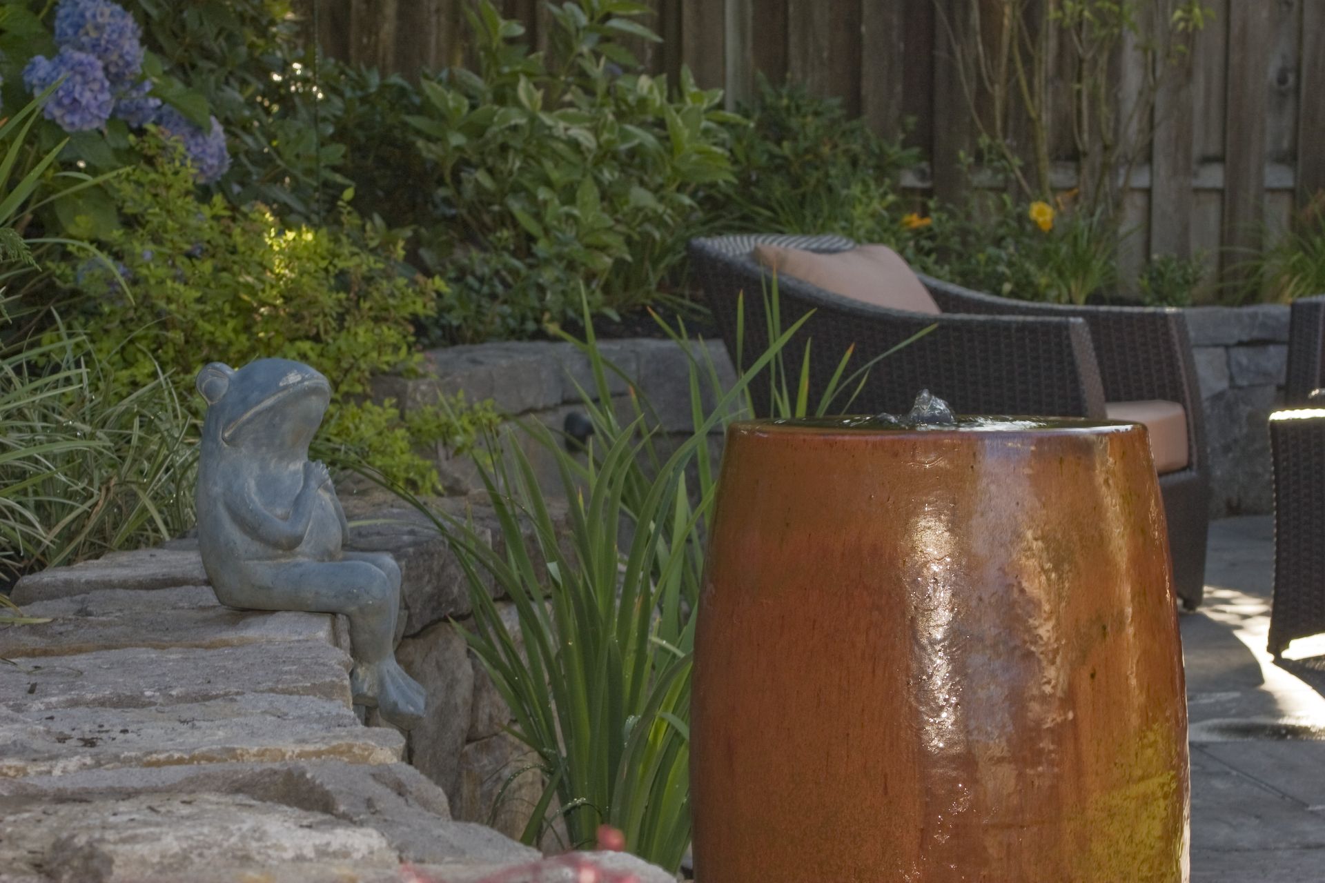 Garden patio with stone wall, bronze fountain, and black outdoor chairs among greenery