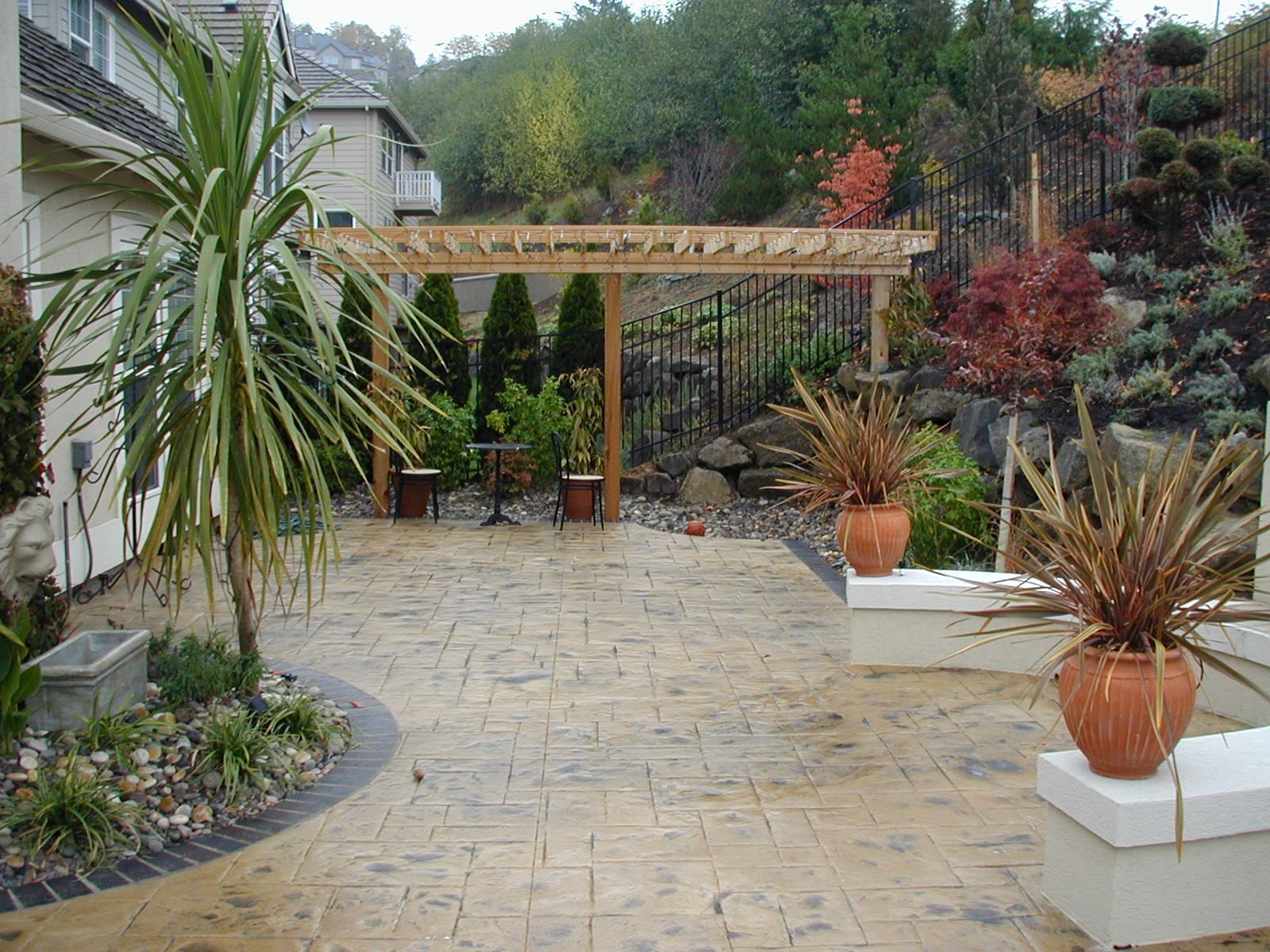 Paved garden patio with potted plants, white planters, and a pergola overlooking a landscaped yard