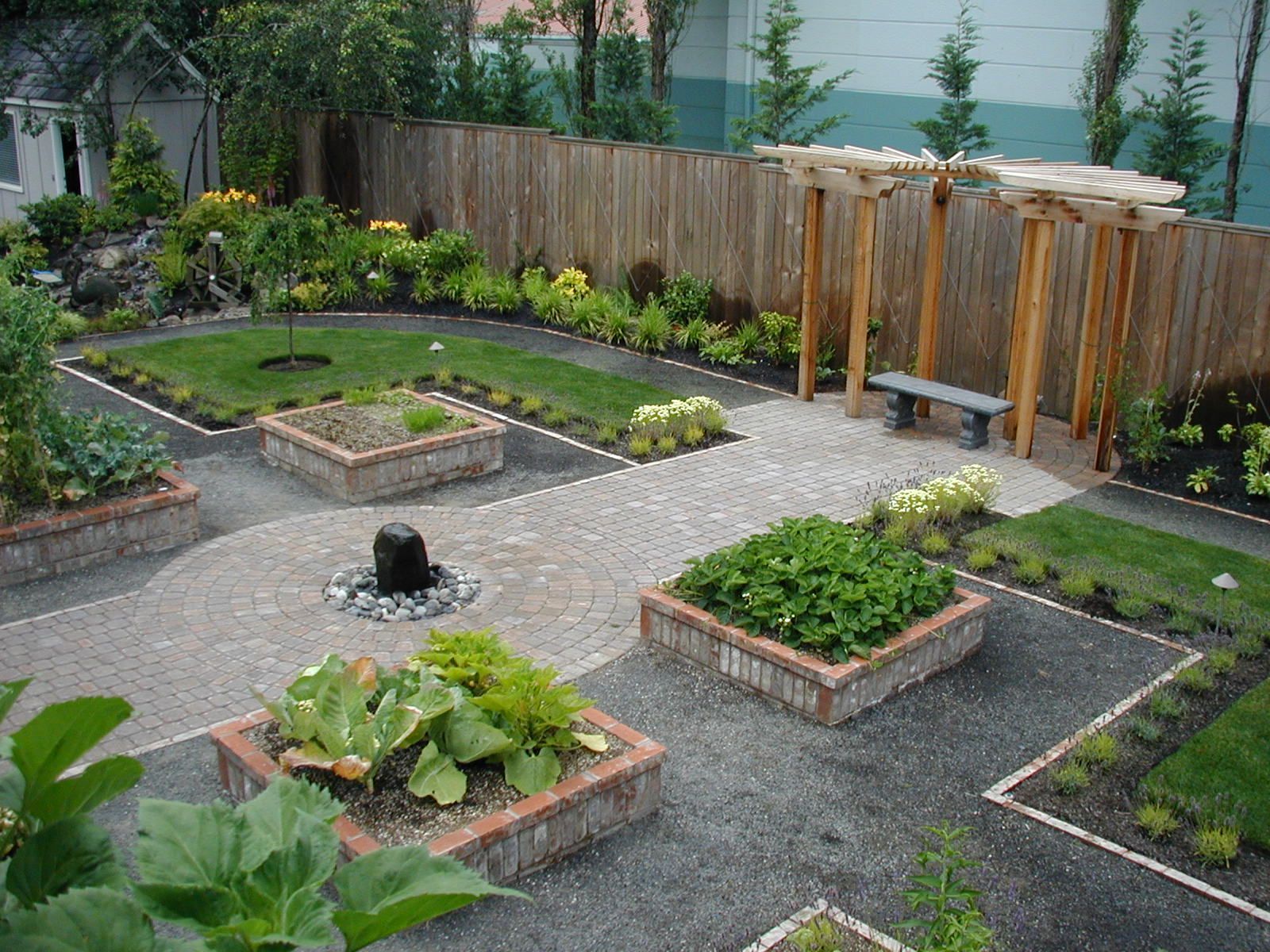 Small backyard garden with raised beds, gray gravel paths, and a wooden fence with benches.