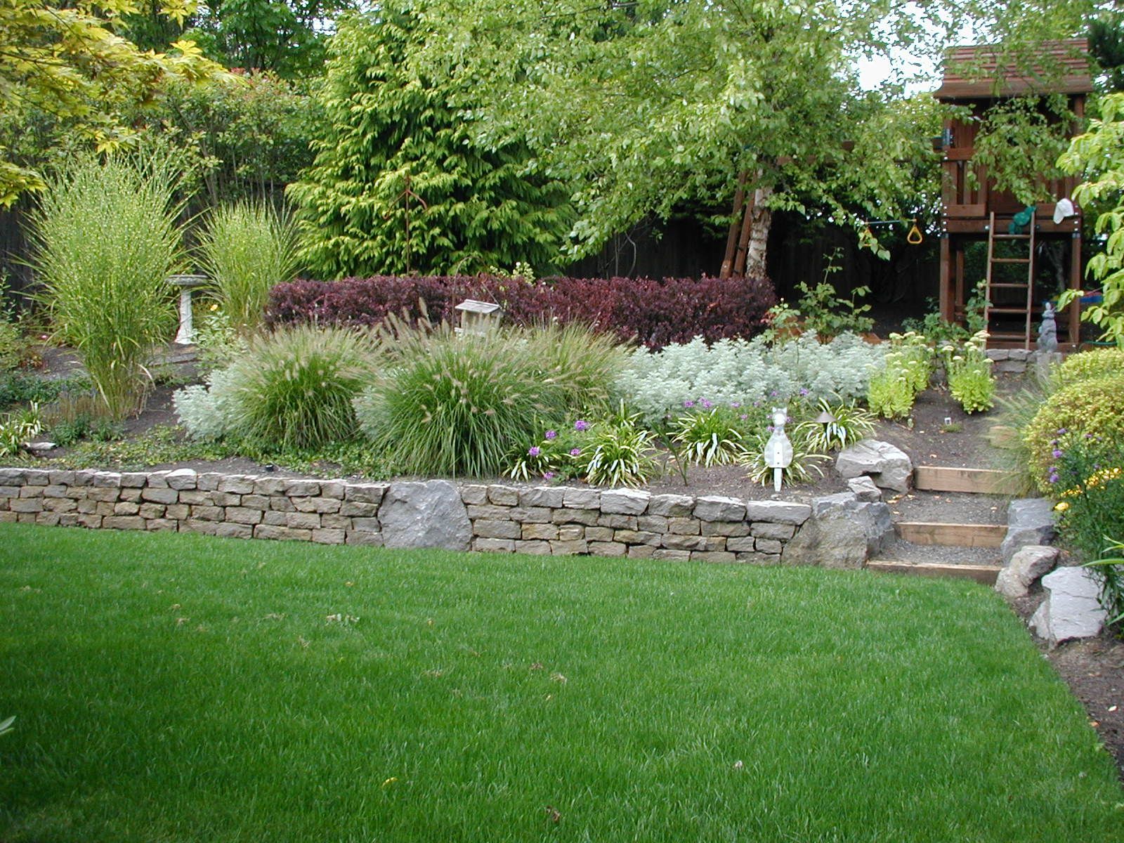 Lush backyard garden with stone retaining wall, white flowers, green lawn, and patio steps under trees