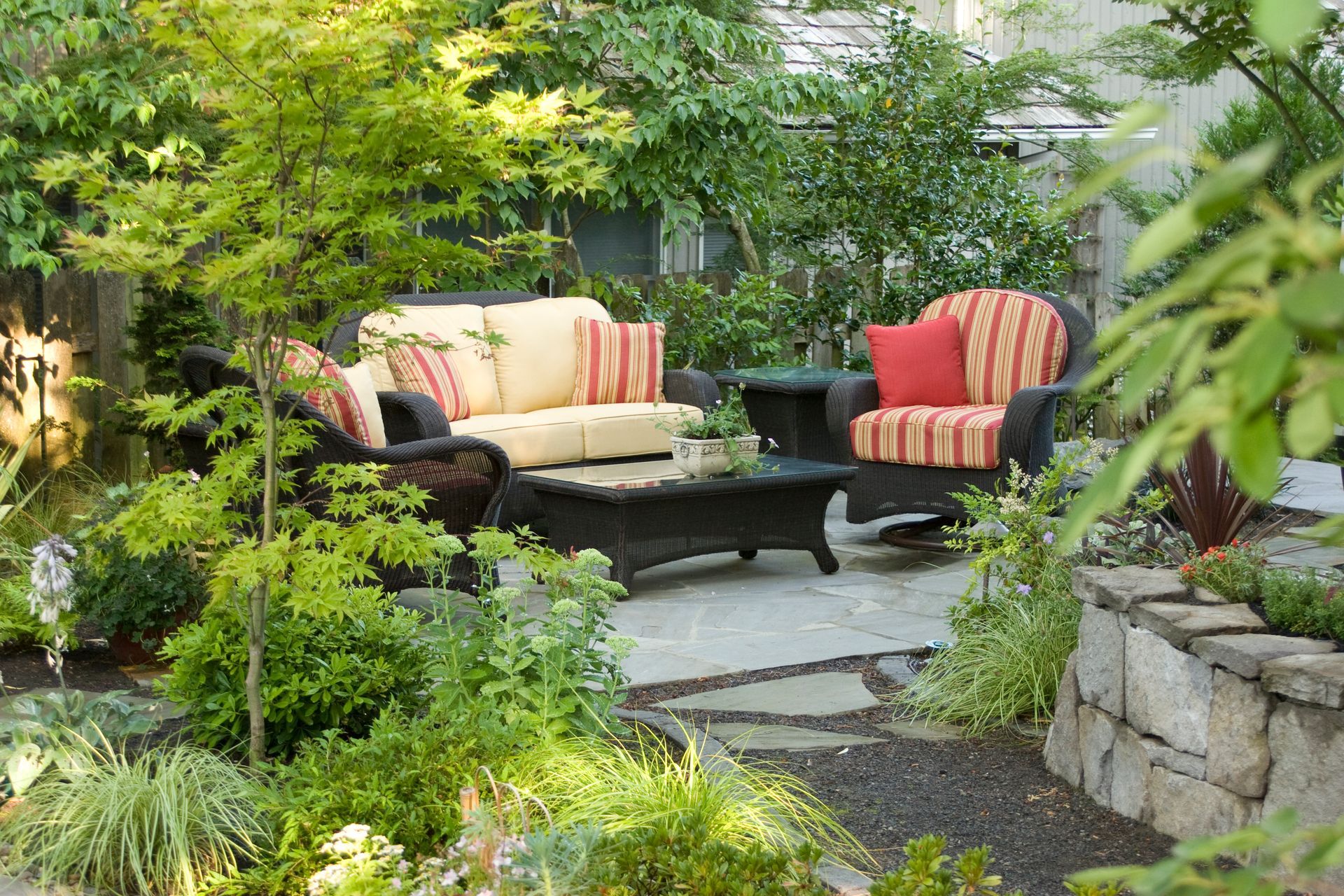 Garden patio with cushioned chairs and a sofa beside lush greenery and stone landscaping
