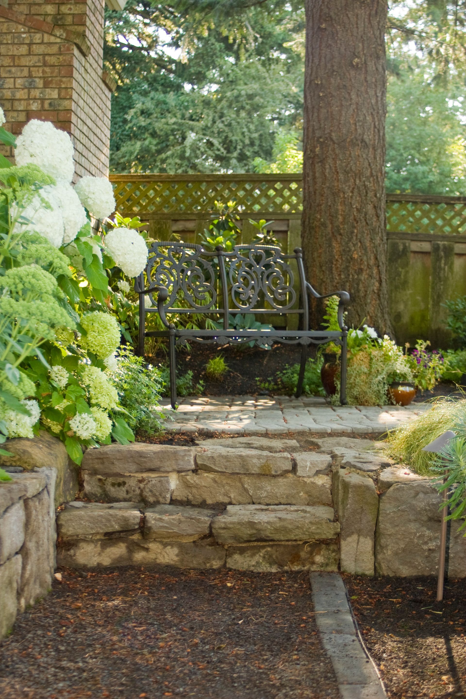 Stone garden steps leading to a black metal bench in a shaded backyard patio surrounded by greenery