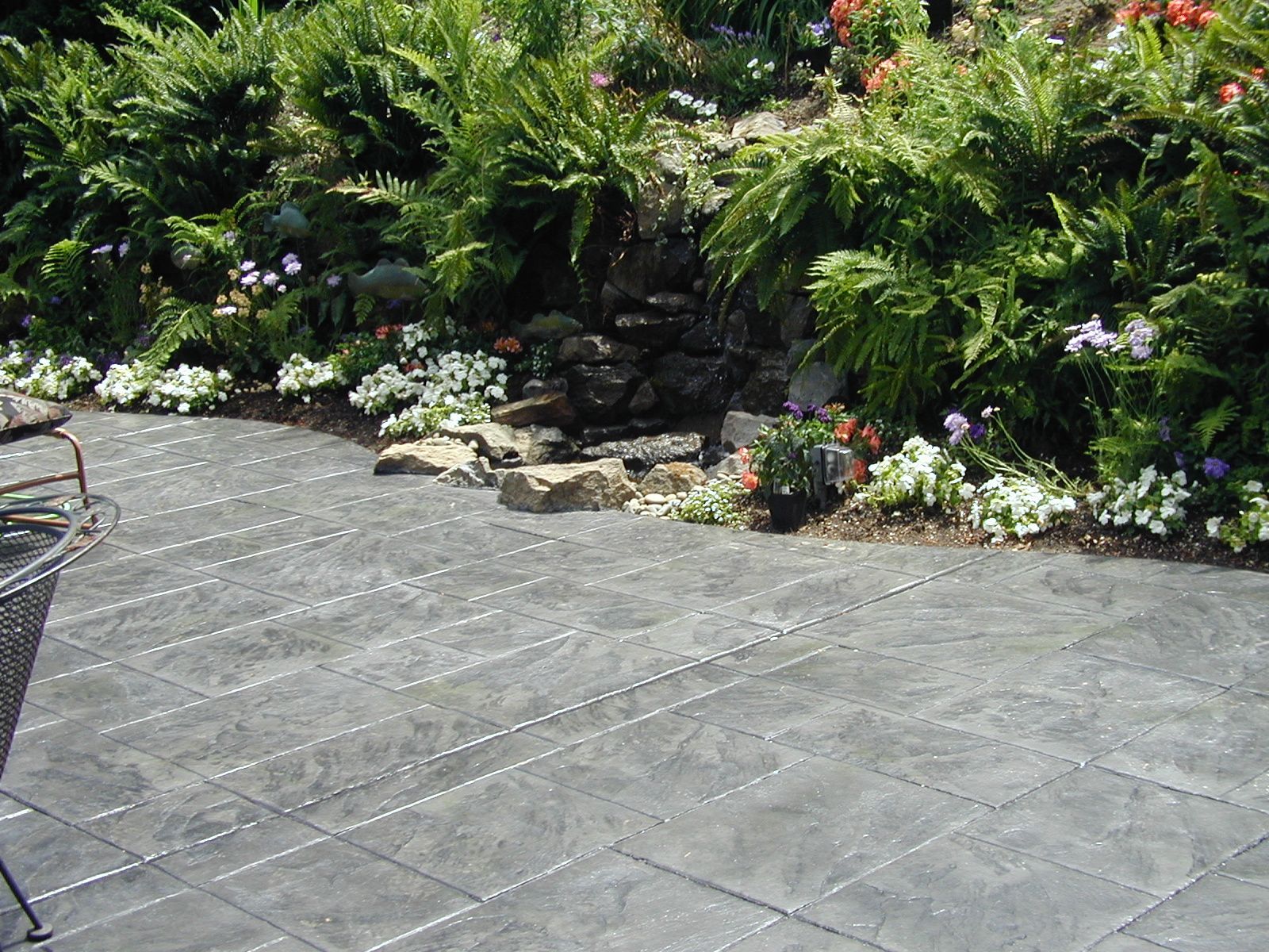 Stone patio beside lush green garden with flowering plants and a wicker chair edge.