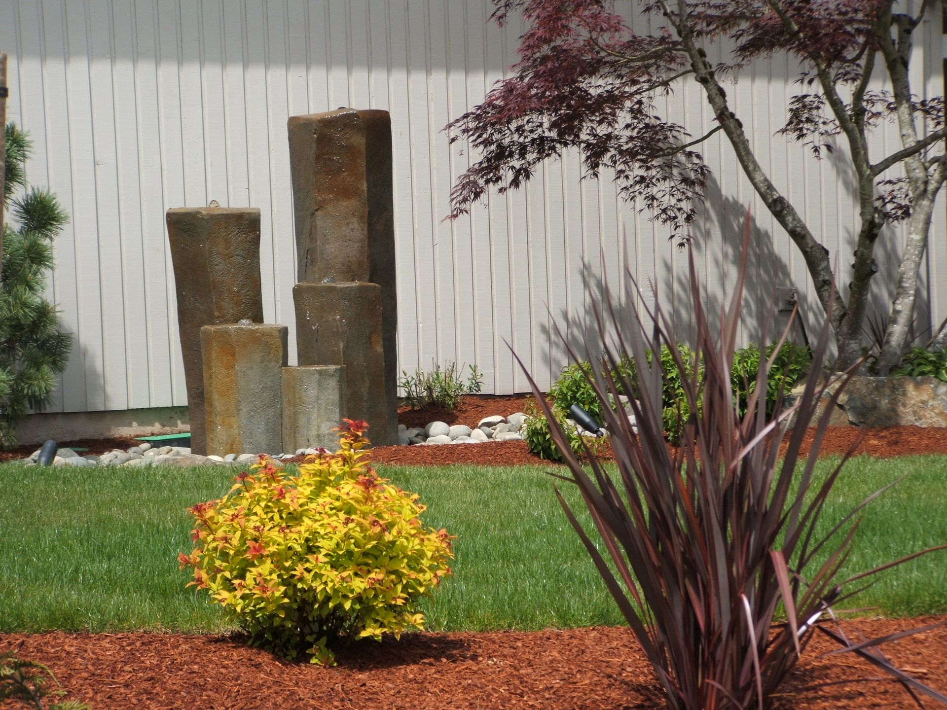 Backyard garden with stacked stone columns, a red-leaf tree, and shrubs against a white fence