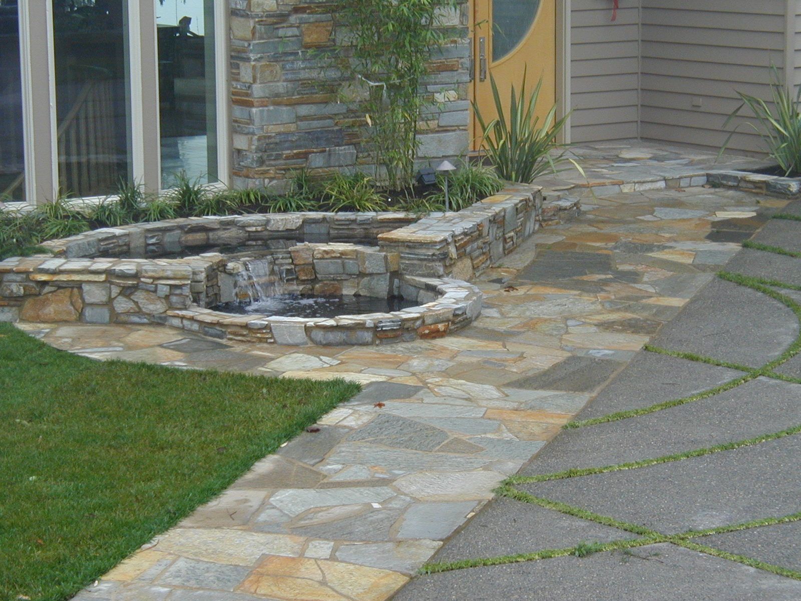 Stone patio and curved retaining wall beside a house, with walkway and green lawn.