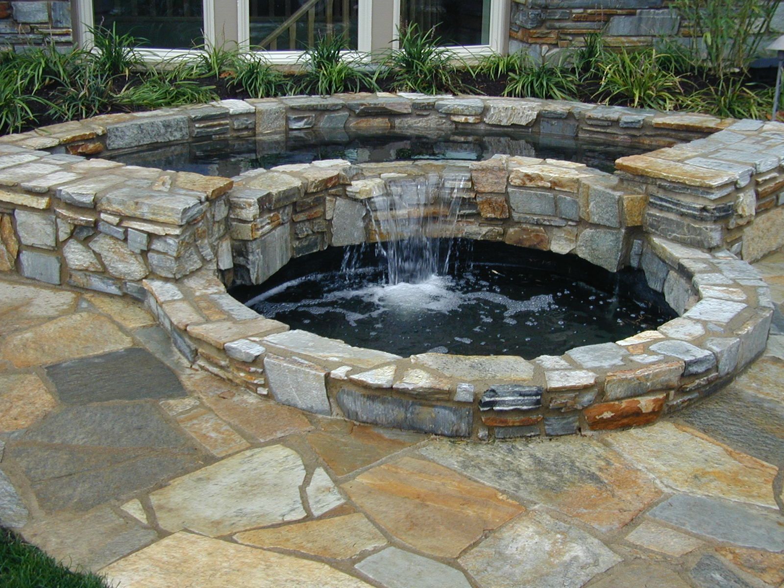 Stone garden fountain with circular black basin and small waterfall, surrounded by flagstone paving.