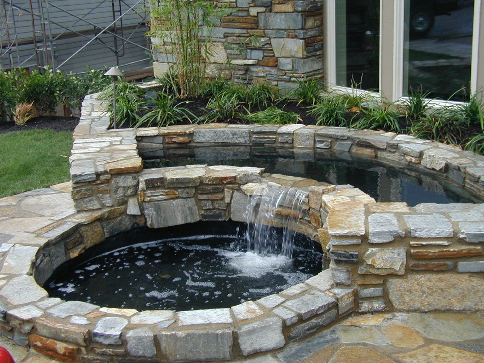Stone backyard fountain with flowing water beside a house and garden plants