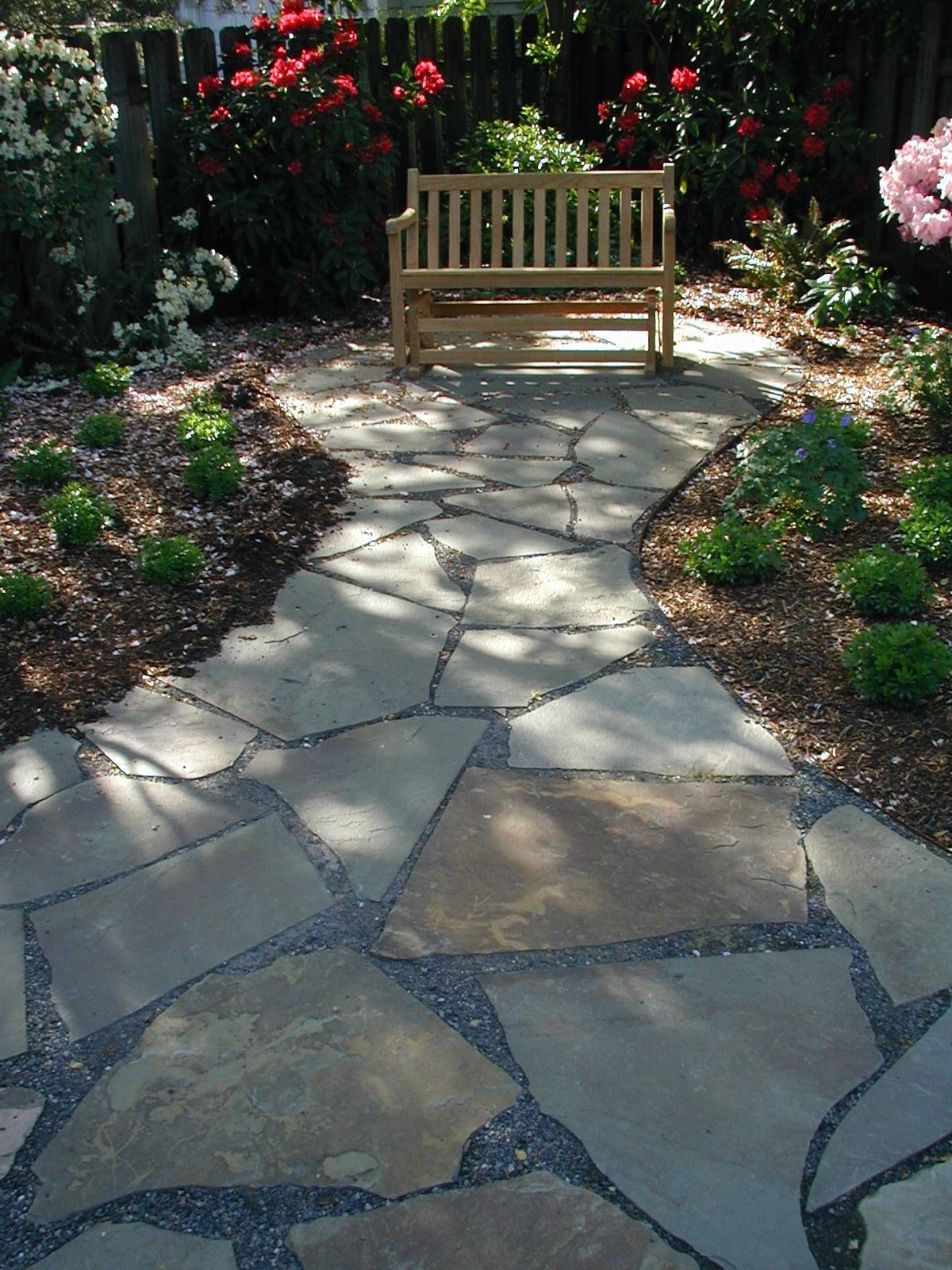 Stone garden path leading to a wooden bench surrounded by flowers and greenery