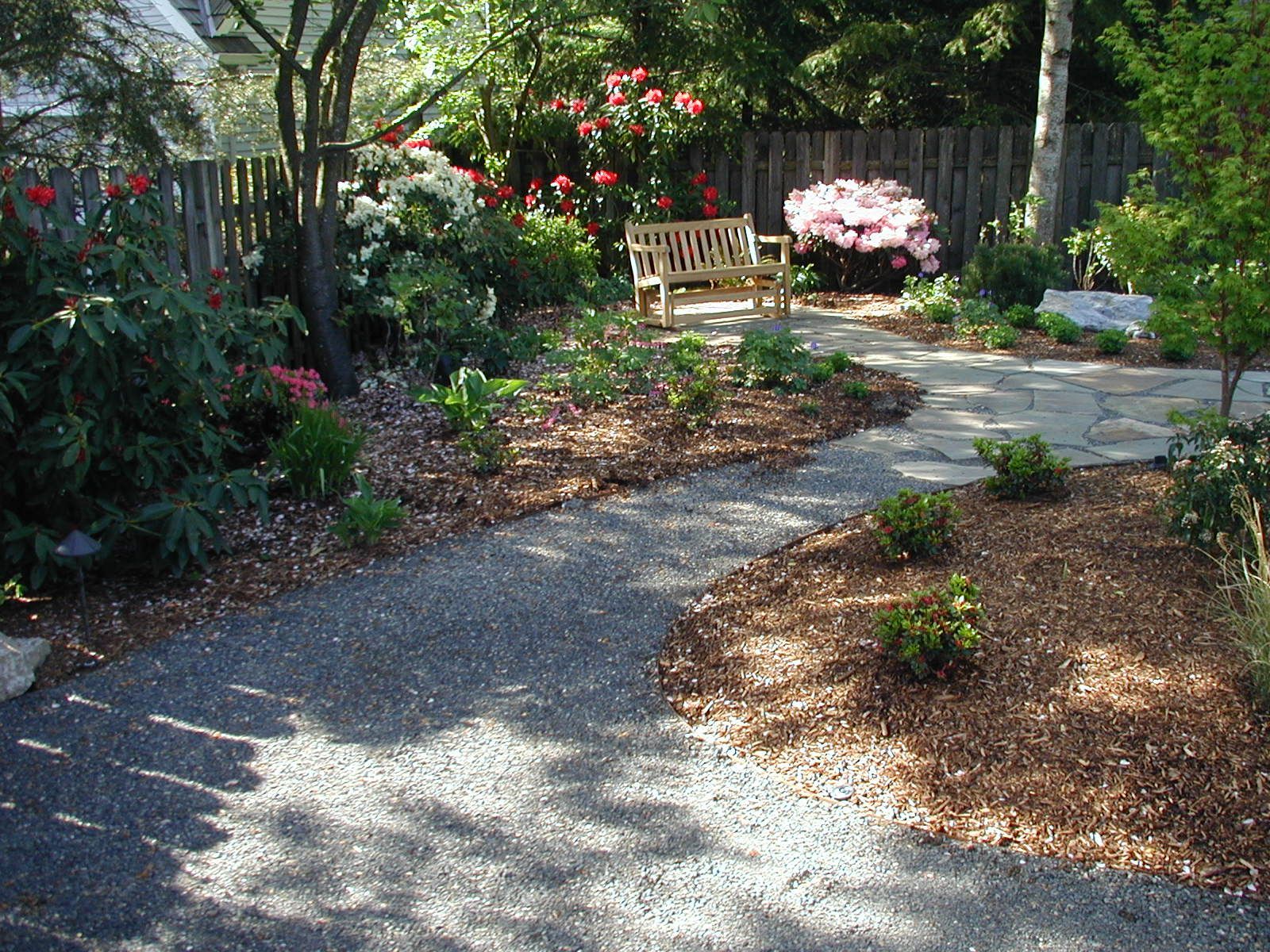 Curved garden path beside flower beds and trees leading to a wooden bench in a sunny yard