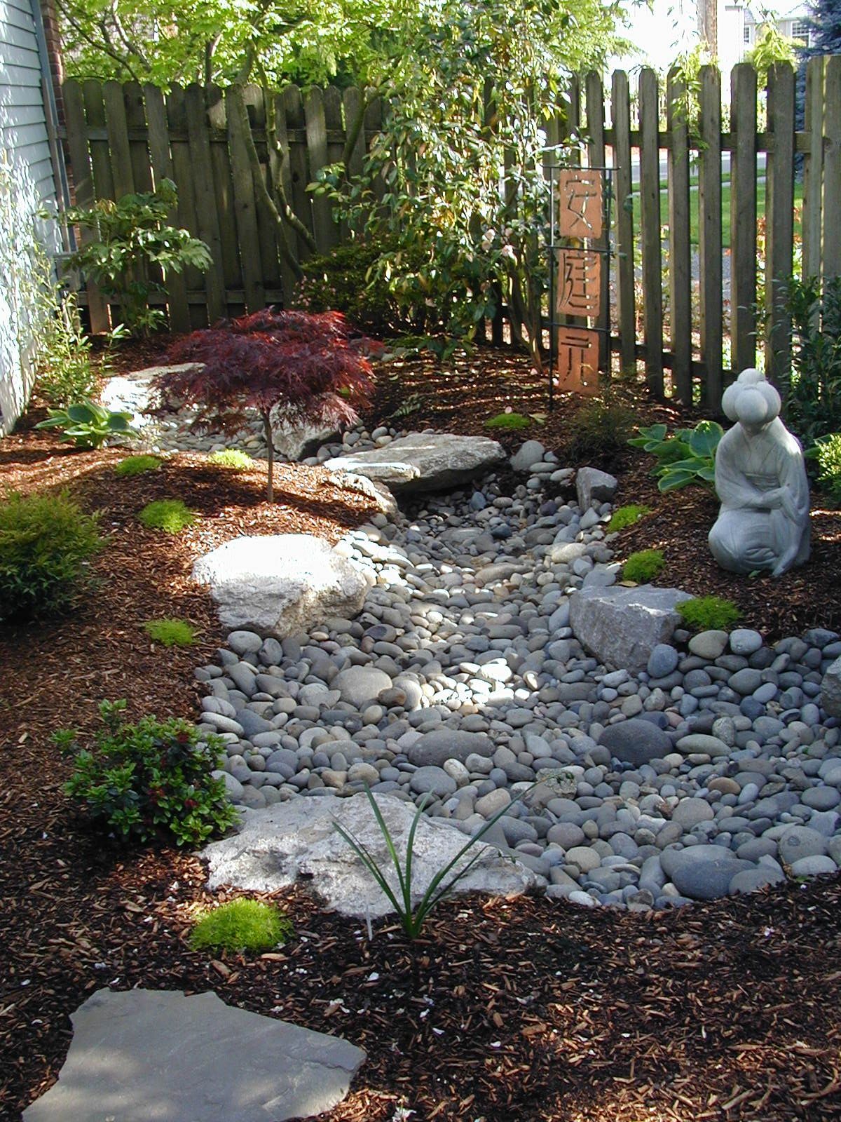Zen garden path with river rocks, stepping stones, a small statue, and a wooden fence in the background