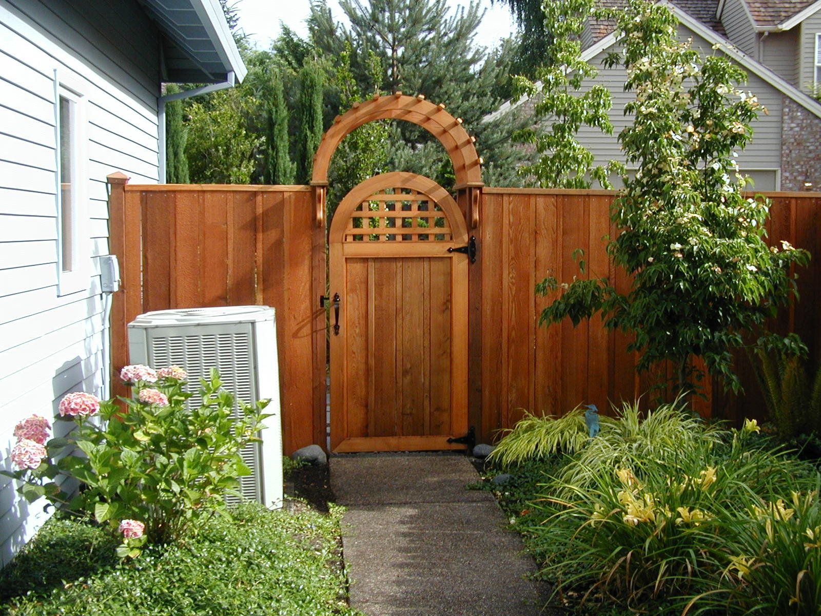 Wooden garden gate with brick arch in a fenced backyard beside a house and walkway