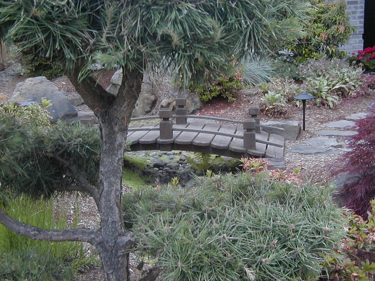 Small arched stone bridge over a garden pond, surrounded by lush trees and shrubs.
