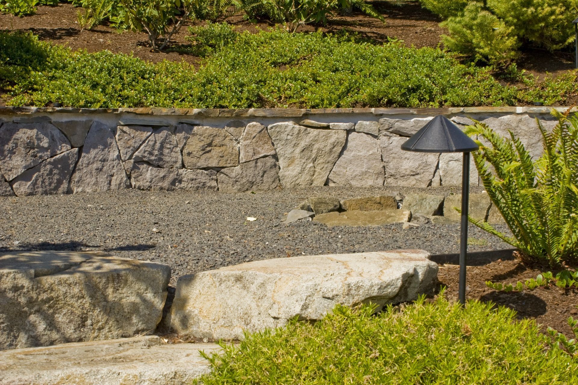 Garden landscape with stone retaining wall, gravel bed, and black path light among green shrubs