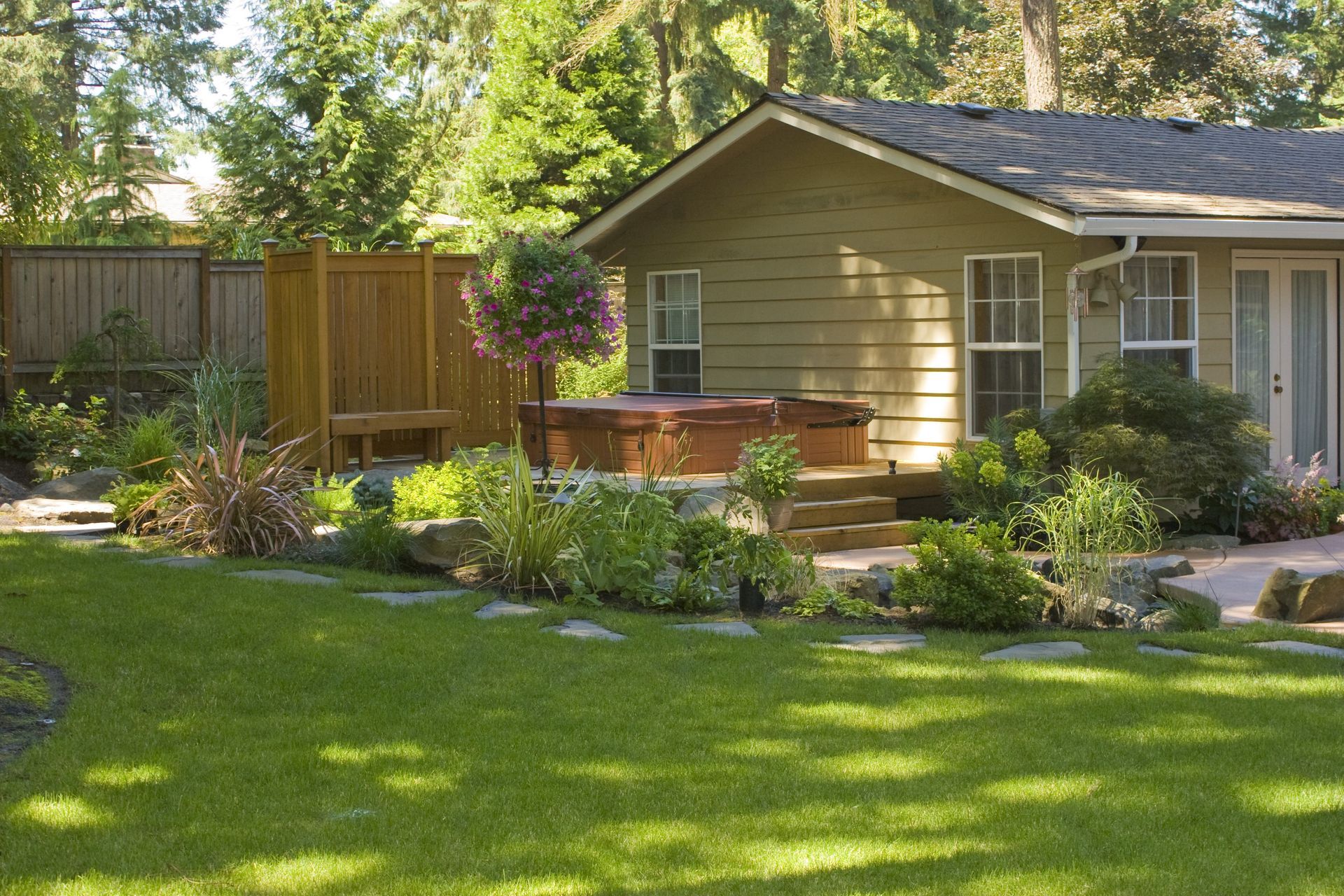 Sunny backyard garden with green lawn, shrubs, wooden deck, and tan house exterior