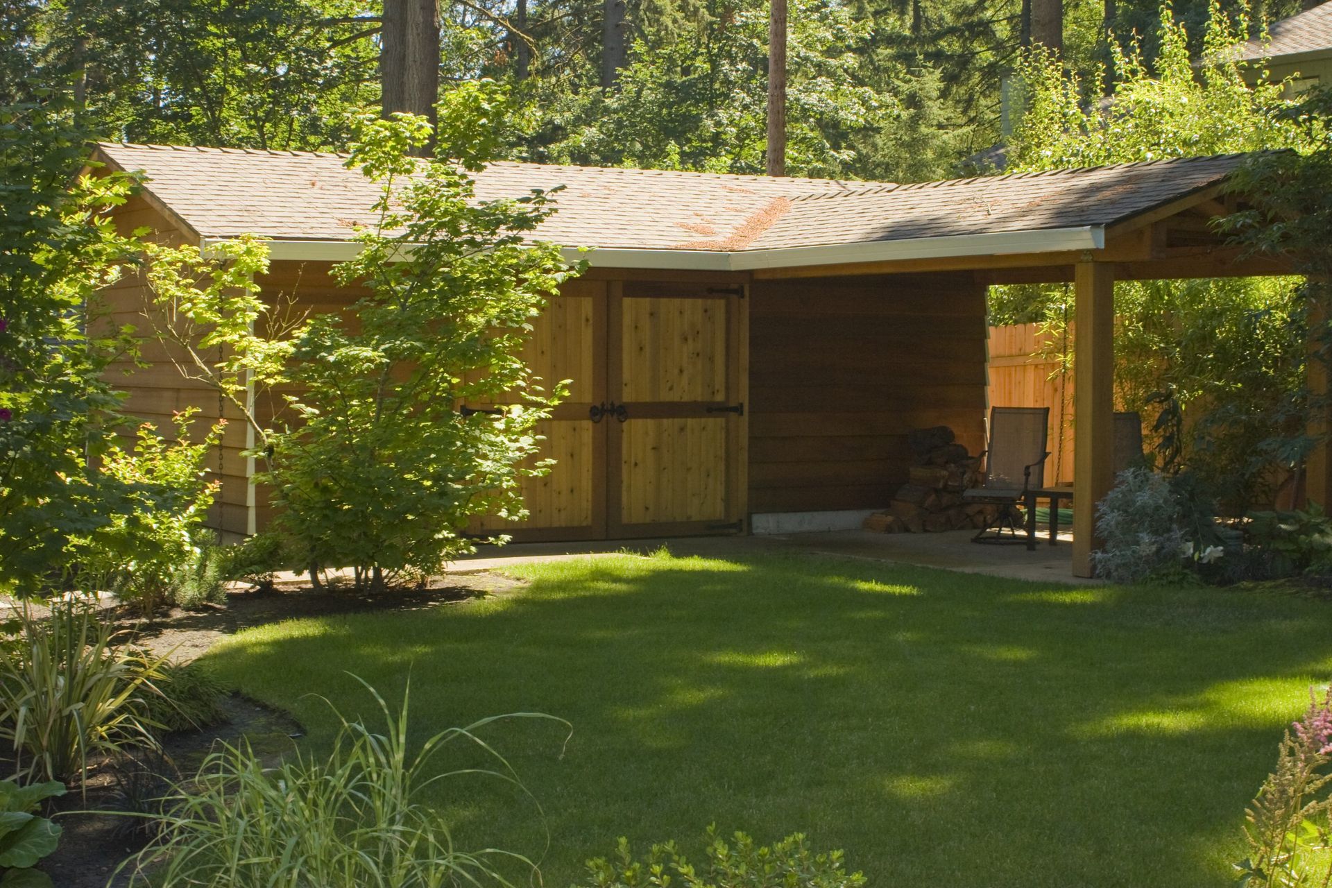 Wooden garden shed with a covered porch in a sunlit green yard, surrounded by trees.