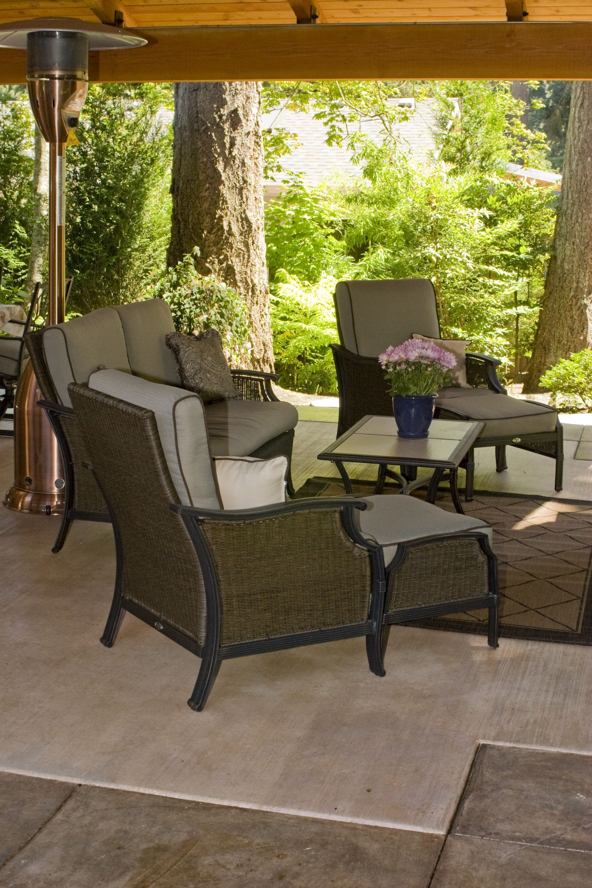 Covered patio with wicker chairs, a small table, and potted flowers in a wooded garden setting